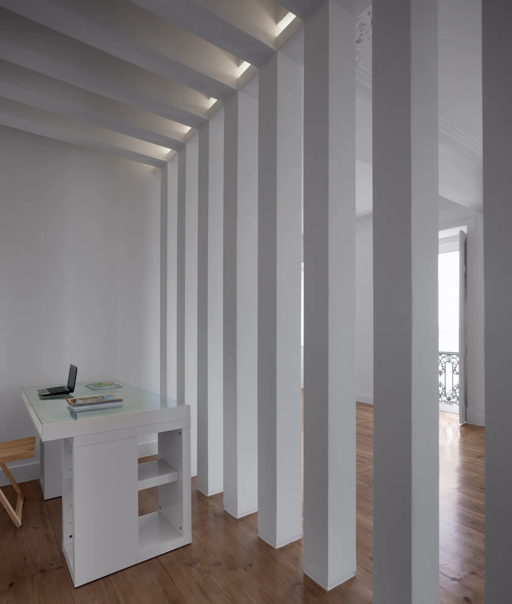 A minimalist home office features a white desk on hardwood floors, separated from an adjacent room by vertical white columns with recessed ceiling lights.