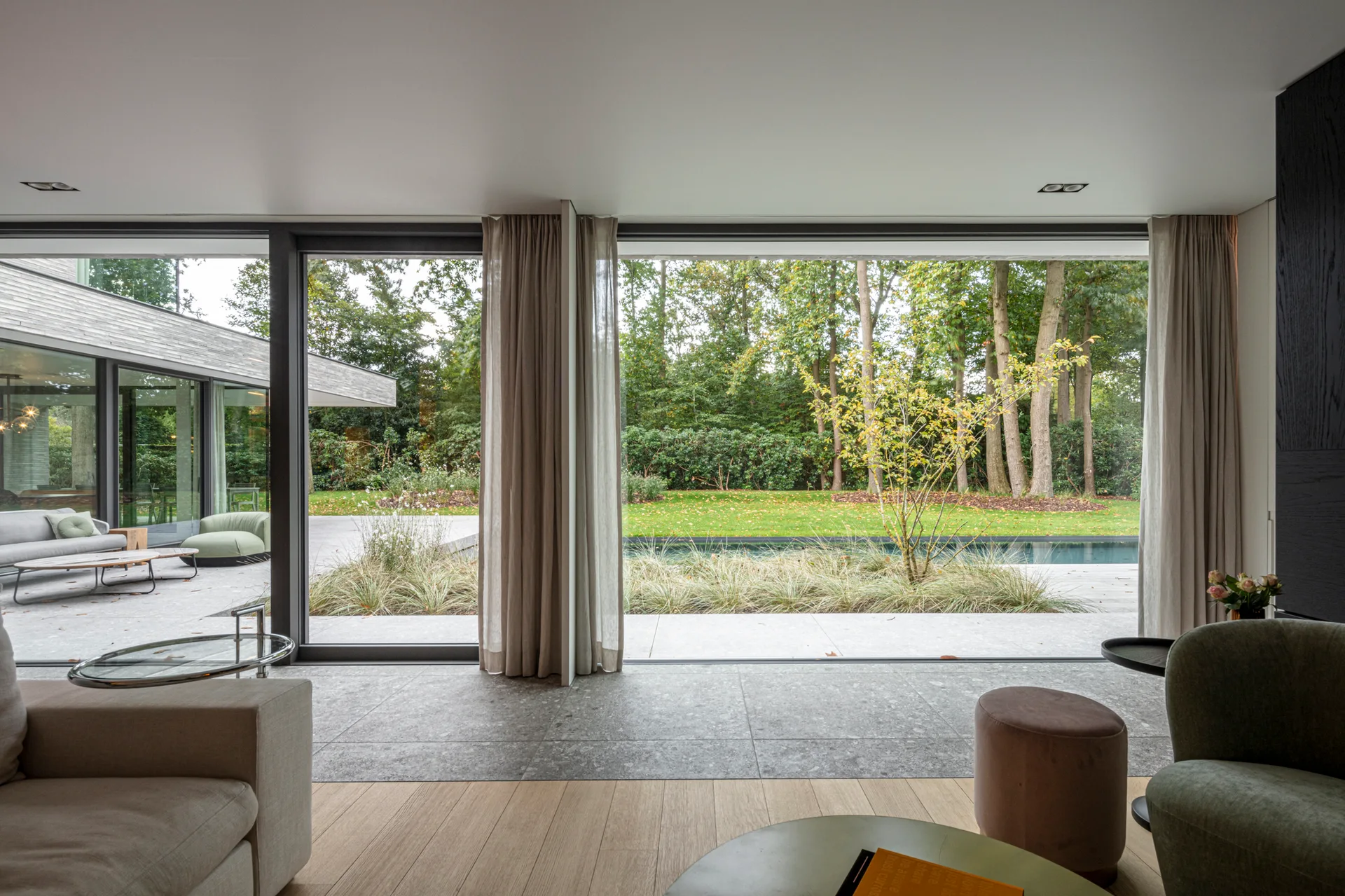 A contemporary living room features floor-to-ceiling glass windows overlooking a garden pool, with stone and wood flooring and sheer curtains.