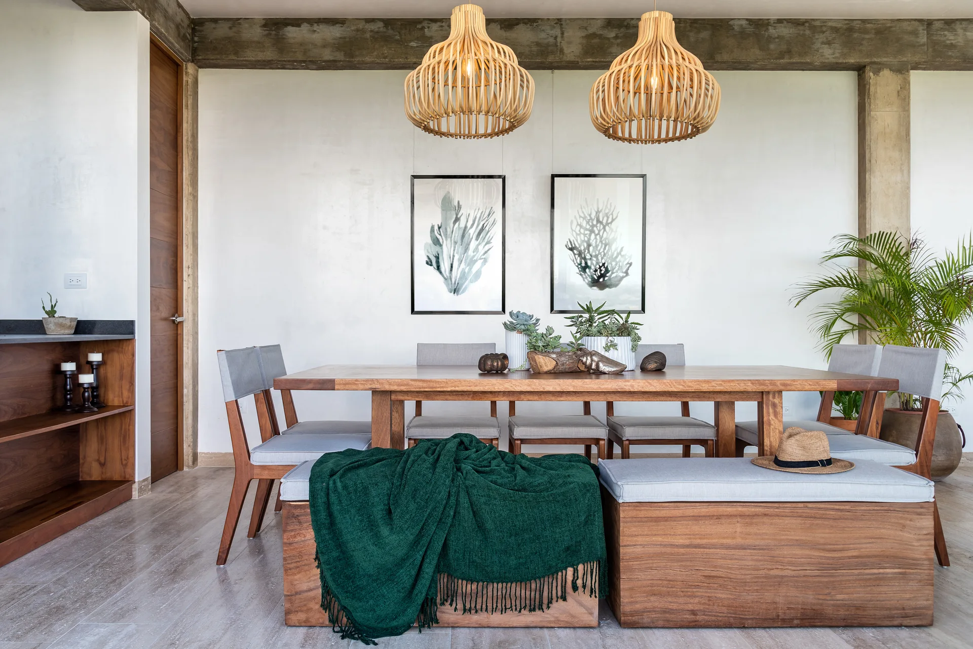 A modern dining room features a wooden table, grey chairs, a cushioned wooden bench with a green throw, and two slatted wooden pendant lights.