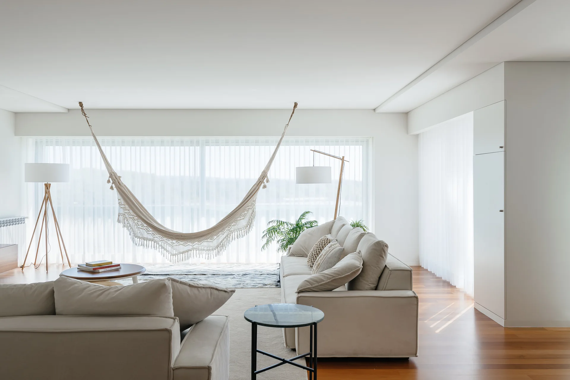 A minimalist living room featuring cream-colored sofas, a woven hammock hung in front of a large window with sheer white curtains, and wood flooring.