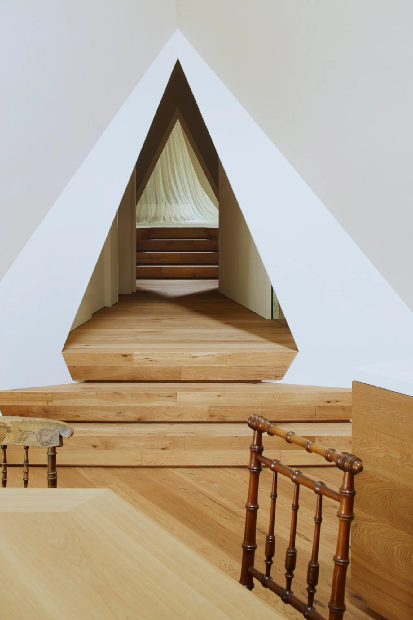 A sharp triangular opening in a white wall frames wooden steps leading up to an adjacent room with sheer white curtains.
