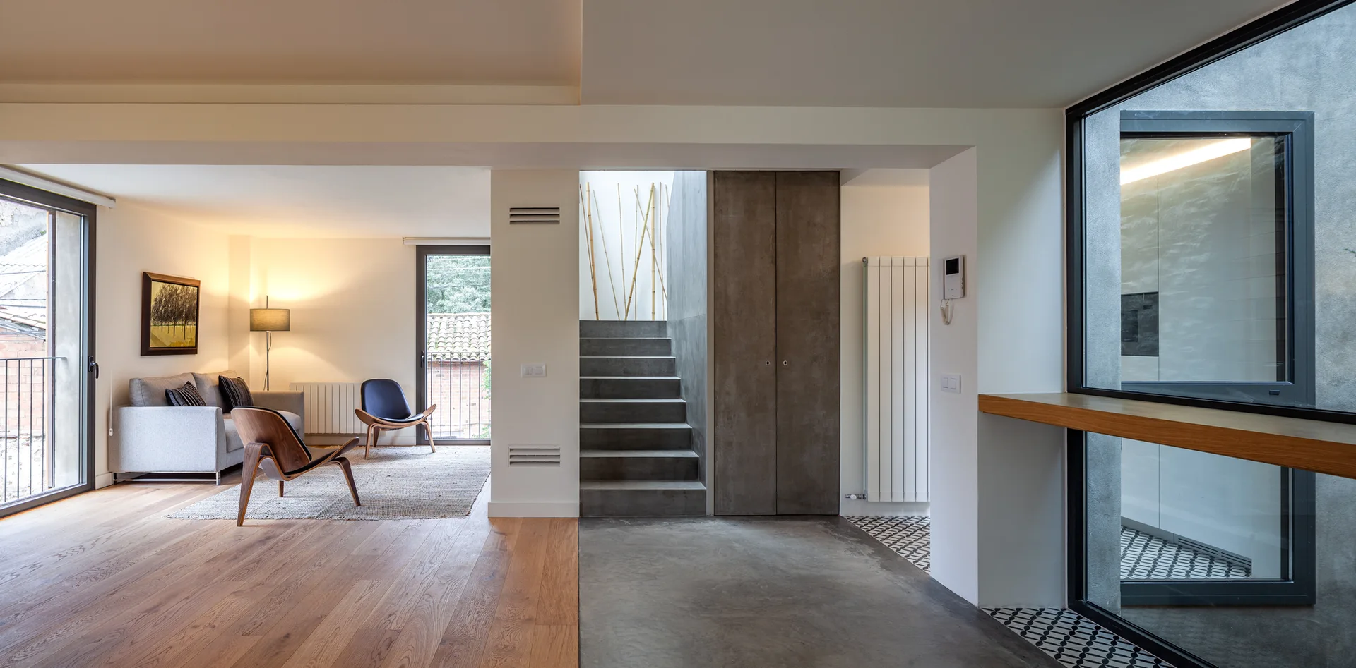 A modern open-plan interior featuring a living room with oak floors, a central concrete staircase, and grey seating next to large windows.