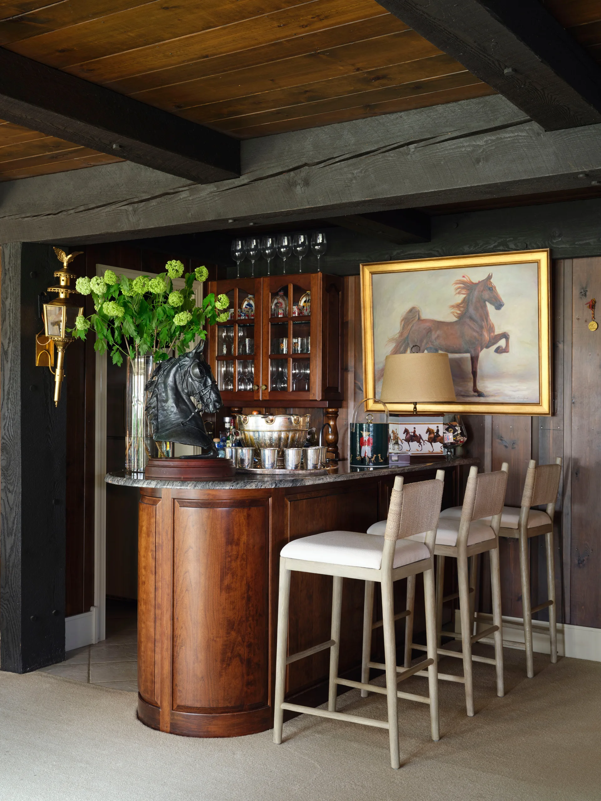 A home bar featuring a curved wood counter, three light-colored upholstered bar stools, and horse-themed artwork on a dark wood-paneled wall.