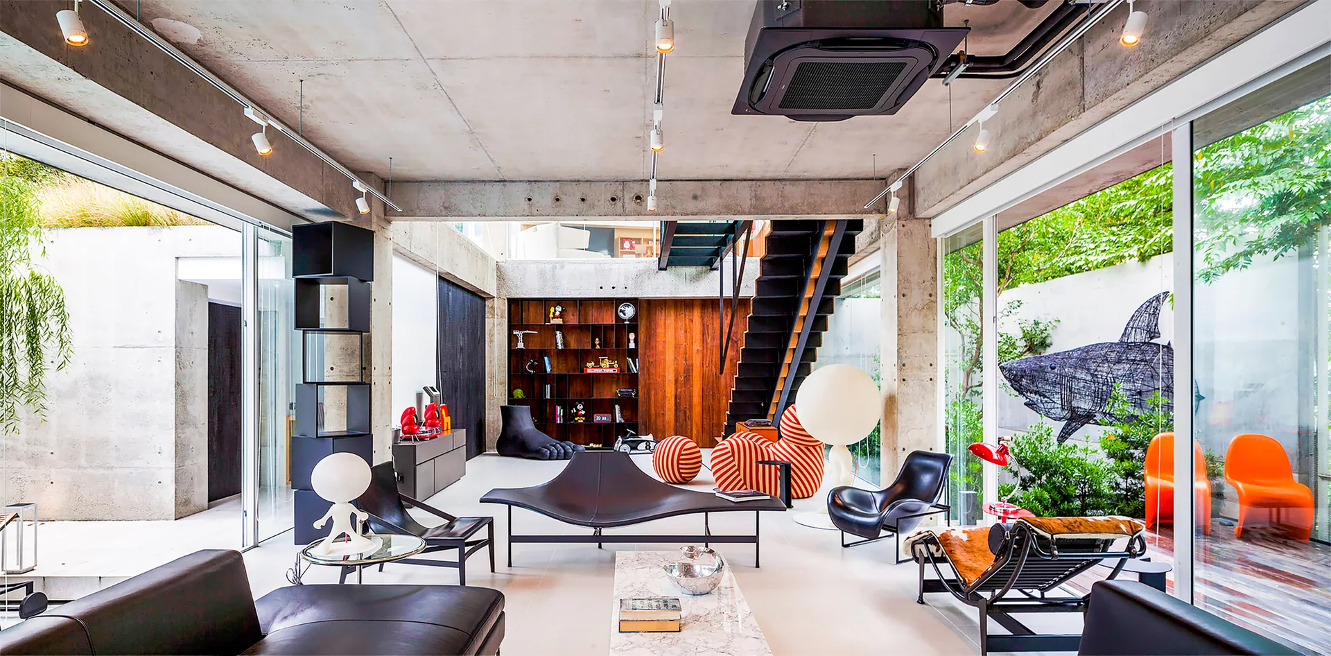 A spacious industrial living room features exposed concrete beams, large glass walls looking onto a garden, and black leather modern furniture.