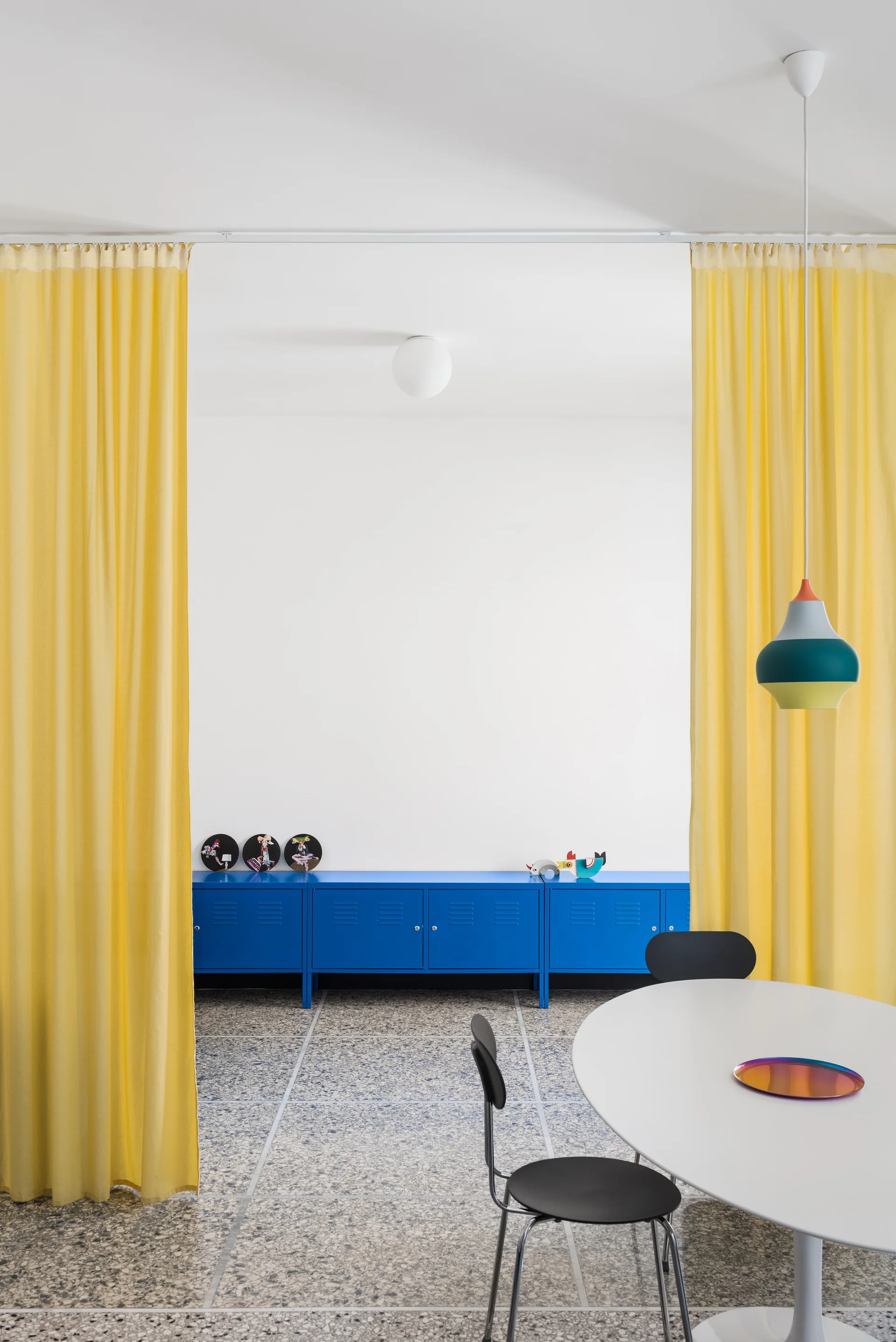 A modern dining room features a white pedestal table, black chairs, a bright blue metal locker sideboard, and yellow curtains.
