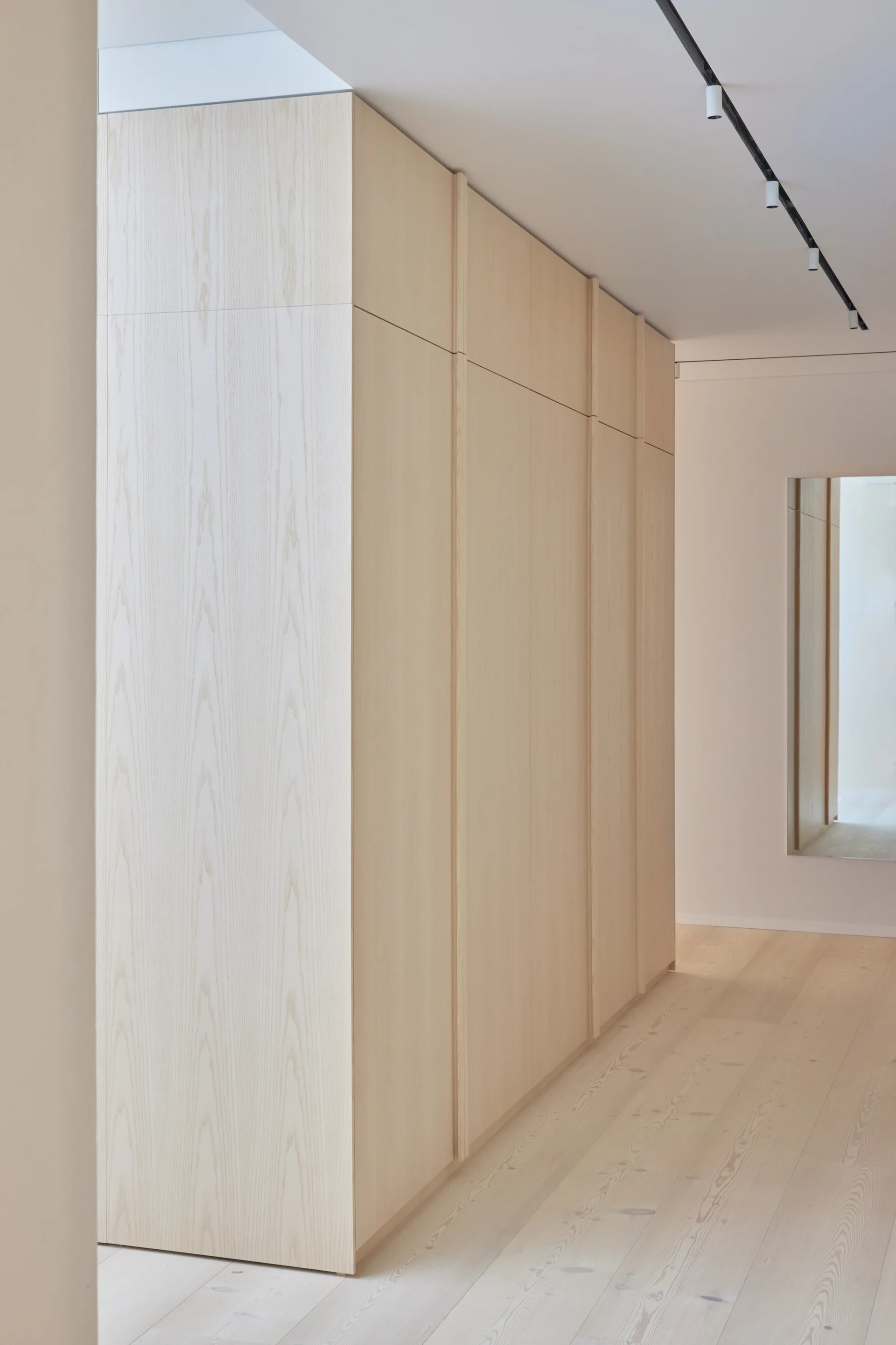 Tall built-in light wood cabinets line a minimalist hallway with matching wide-plank flooring and black ceiling track lighting.