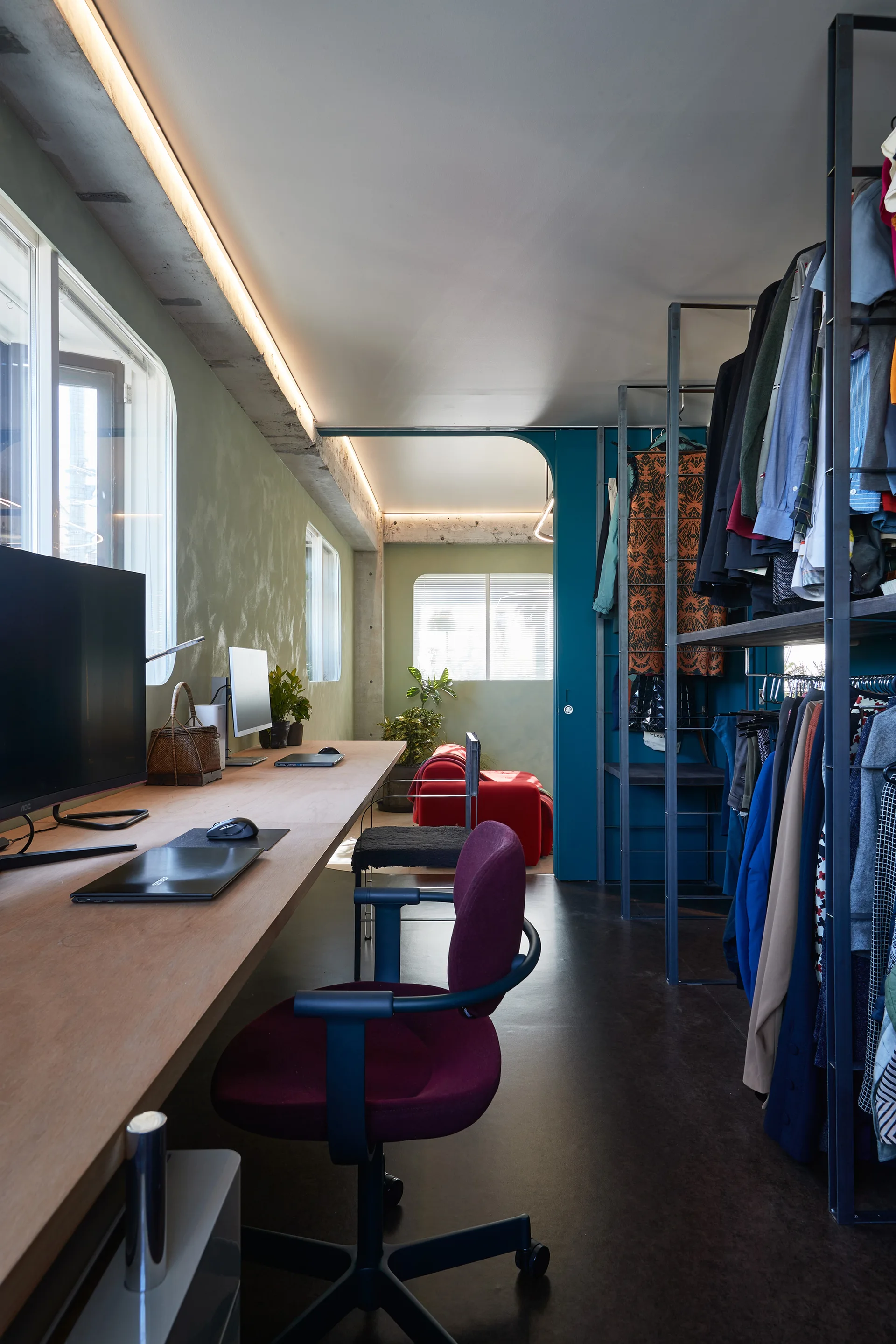 A long wooden desk with computer monitors stands against a textured sage green wall beside an open metal clothing rack.