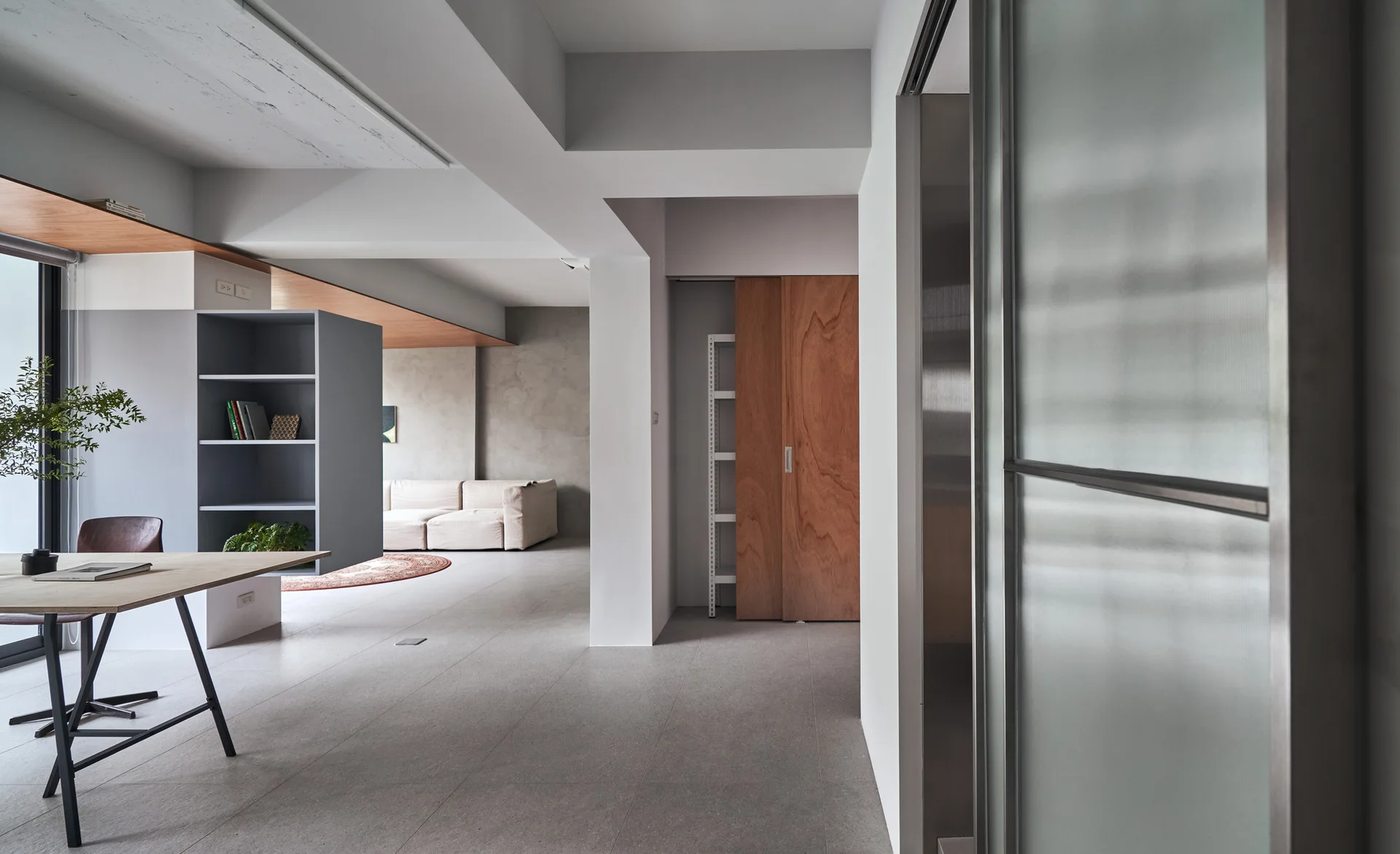 A modern open-plan living room features grey tile floors, a wooden dining table, a grey shelving unit divider, and a beige sofa.