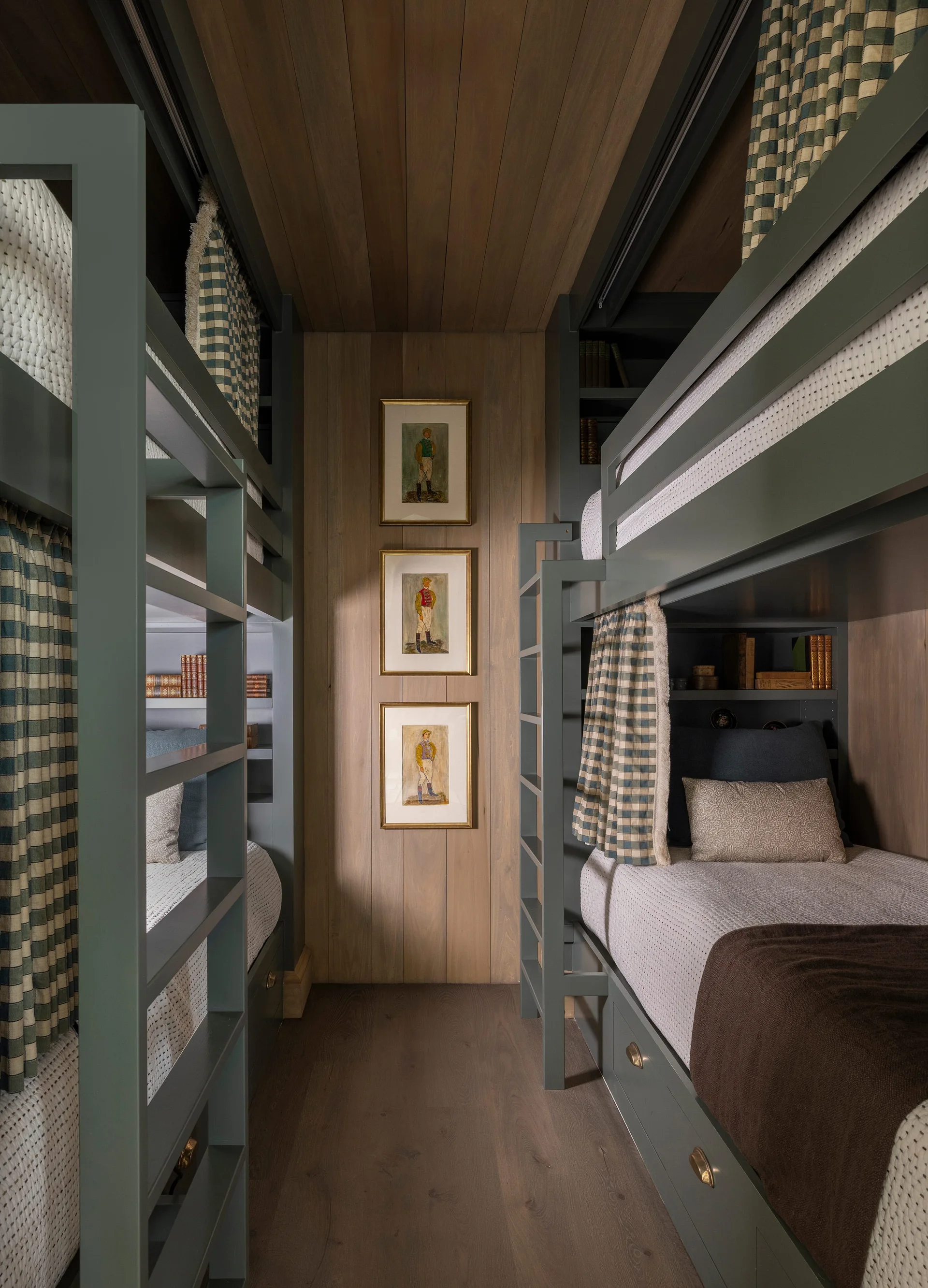 Symmetrical sage green bunk beds built into a narrow room with warm wood-paneled walls, checkered curtains, and stacked framed art.