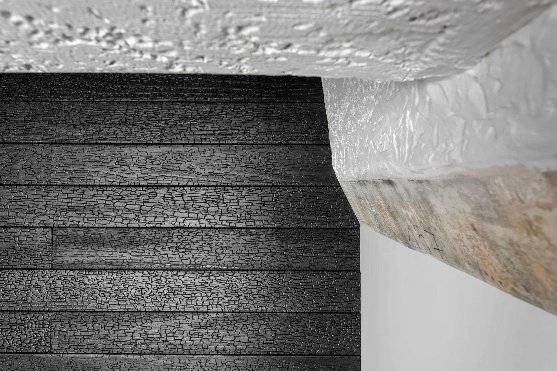A close-up view of dark charred wood wall paneling in a horizontal plank pattern meeting a rough-textured white plaster ceiling beam.