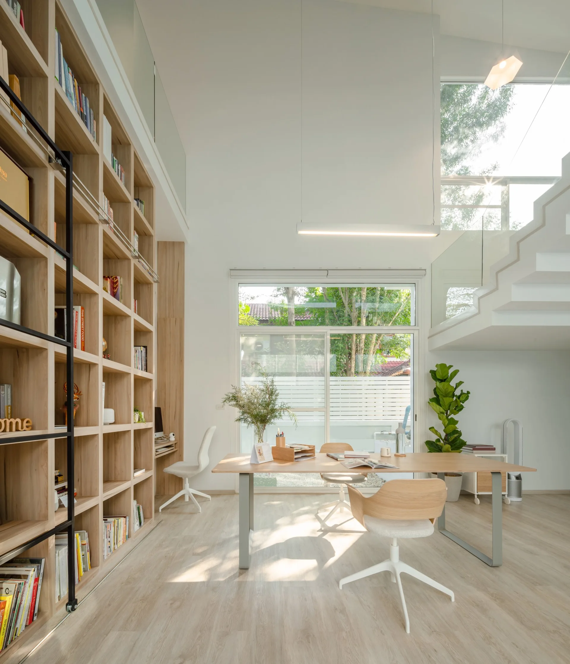 A bright double-height home office features a massive floor-to-ceiling wooden bookshelf, a central desk with modern chairs, and a white staircase.