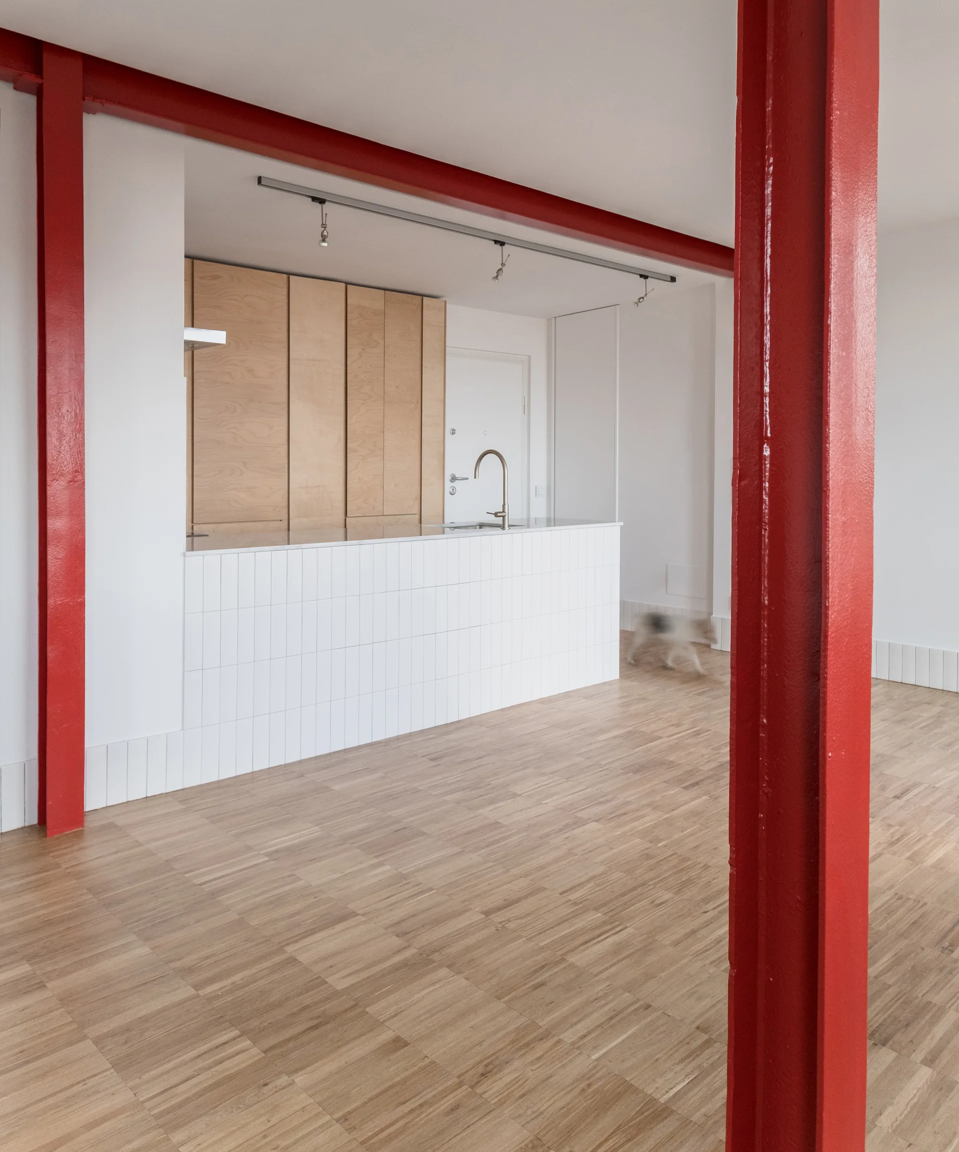 A modern open kitchen features bright red structural steel columns, a vertical-tiled island, flat light wood cabinets, and parquet wood flooring.