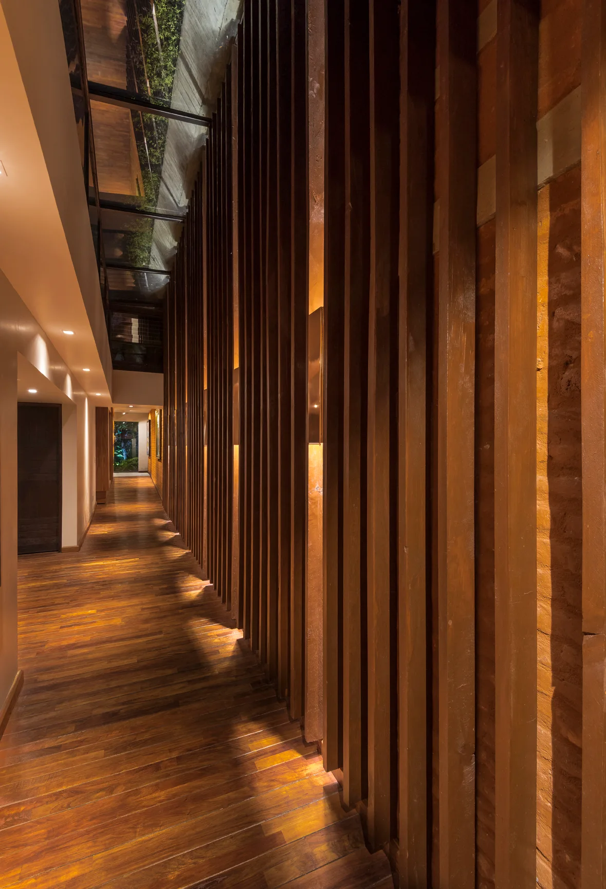 A long hallway with polished hardwood floors is lined by a wall of vertical wooden slats illuminated by integrated warm accent lighting.