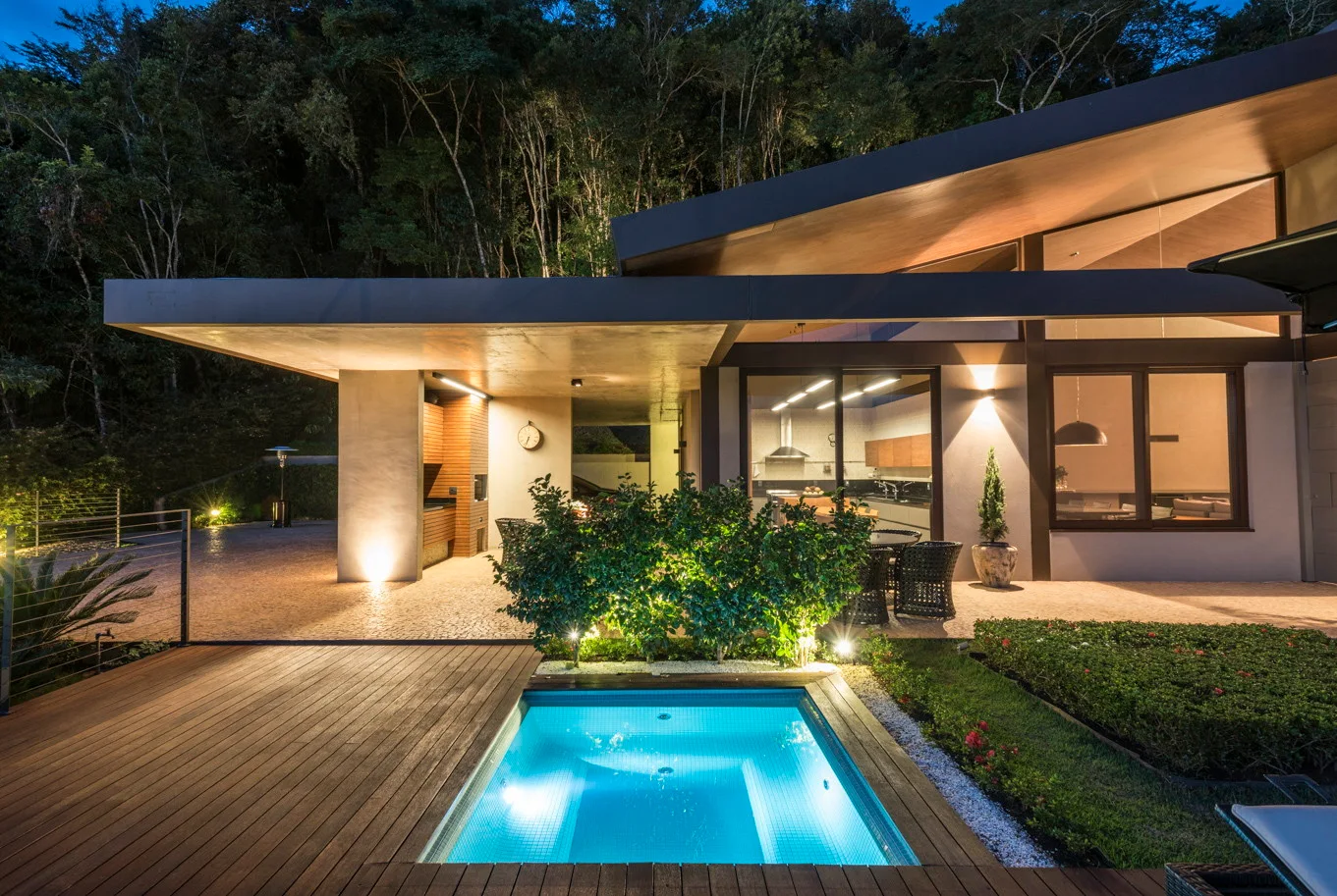 A modern home features an illuminated rectangular pool set into a wood deck next to a covered stone patio at dusk.