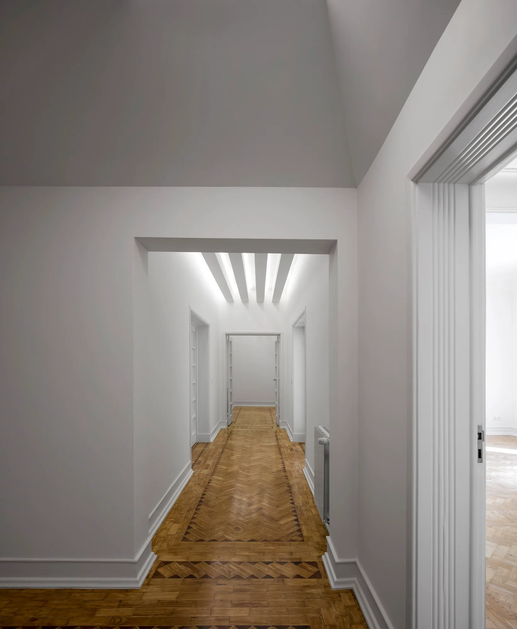A long white residential corridor features intricate herringbone parquet flooring, classic molded doorframes, and ceiling beams.