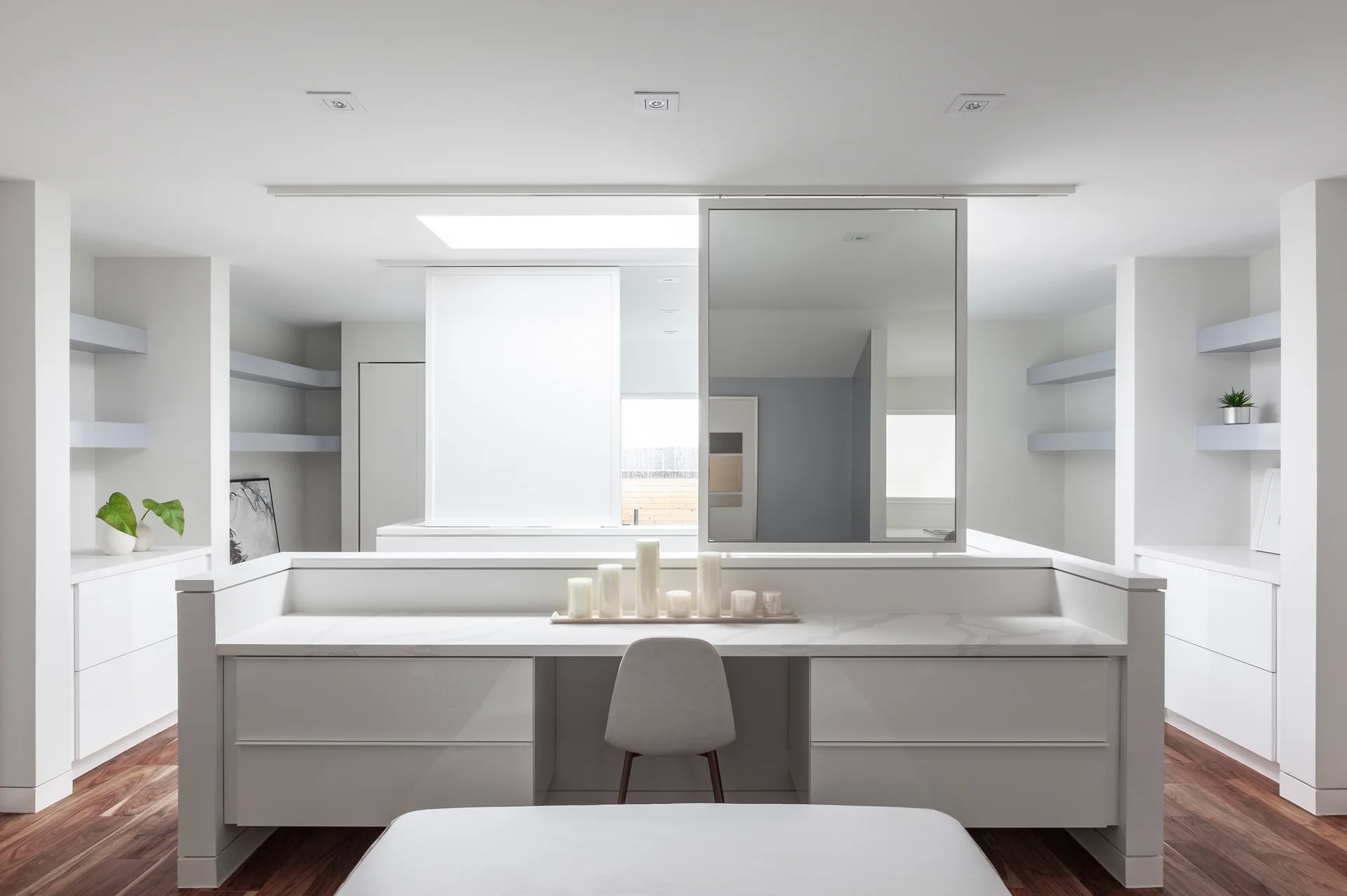 A modern white dressing room features a central vanity island with a marble-patterned countertop, a grey chair, and symmetrical built-in cabinets.
