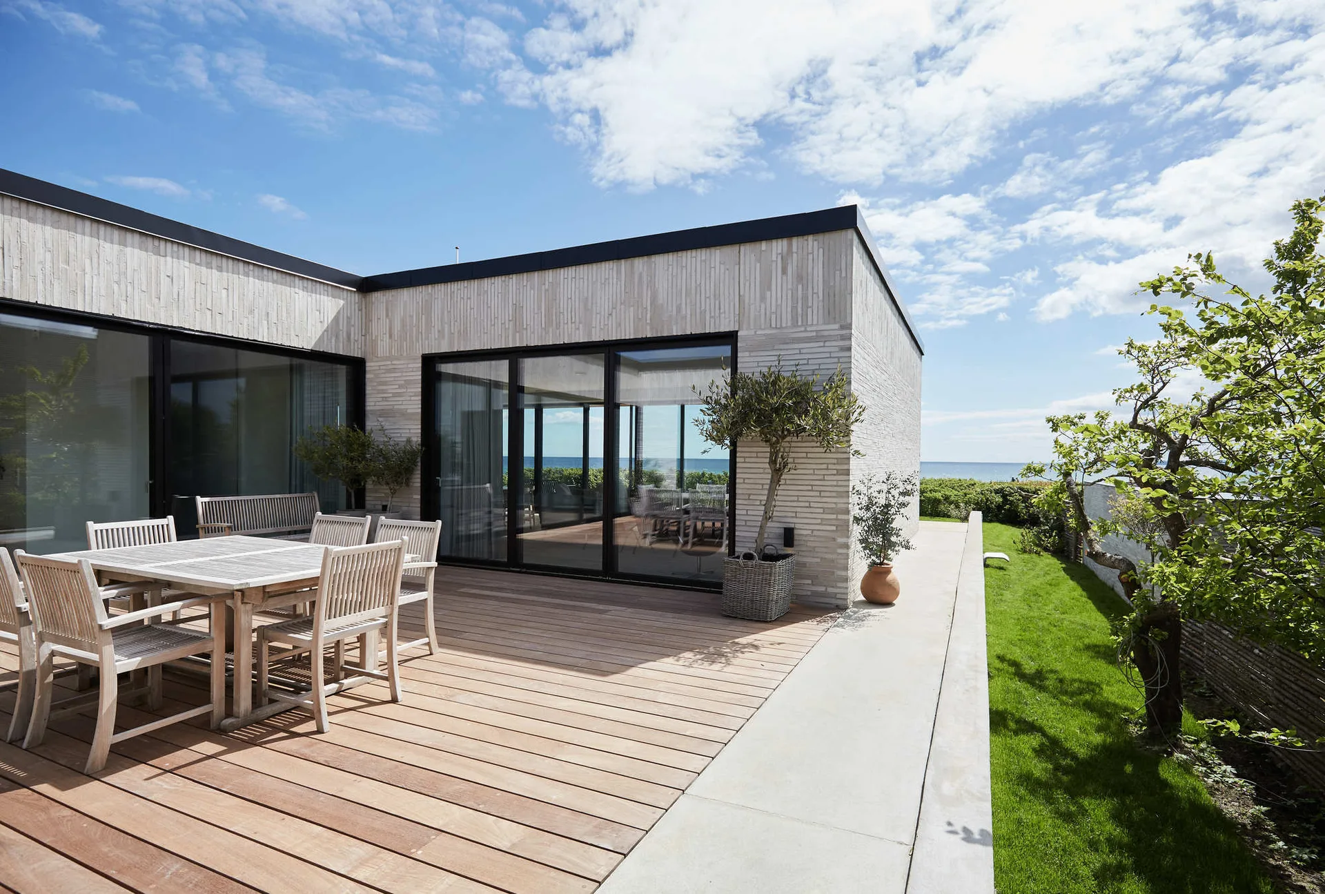 A wooden deck features a slatted wood dining table and chairs next to a modern light-brick house with large black-framed sliding glass doors.