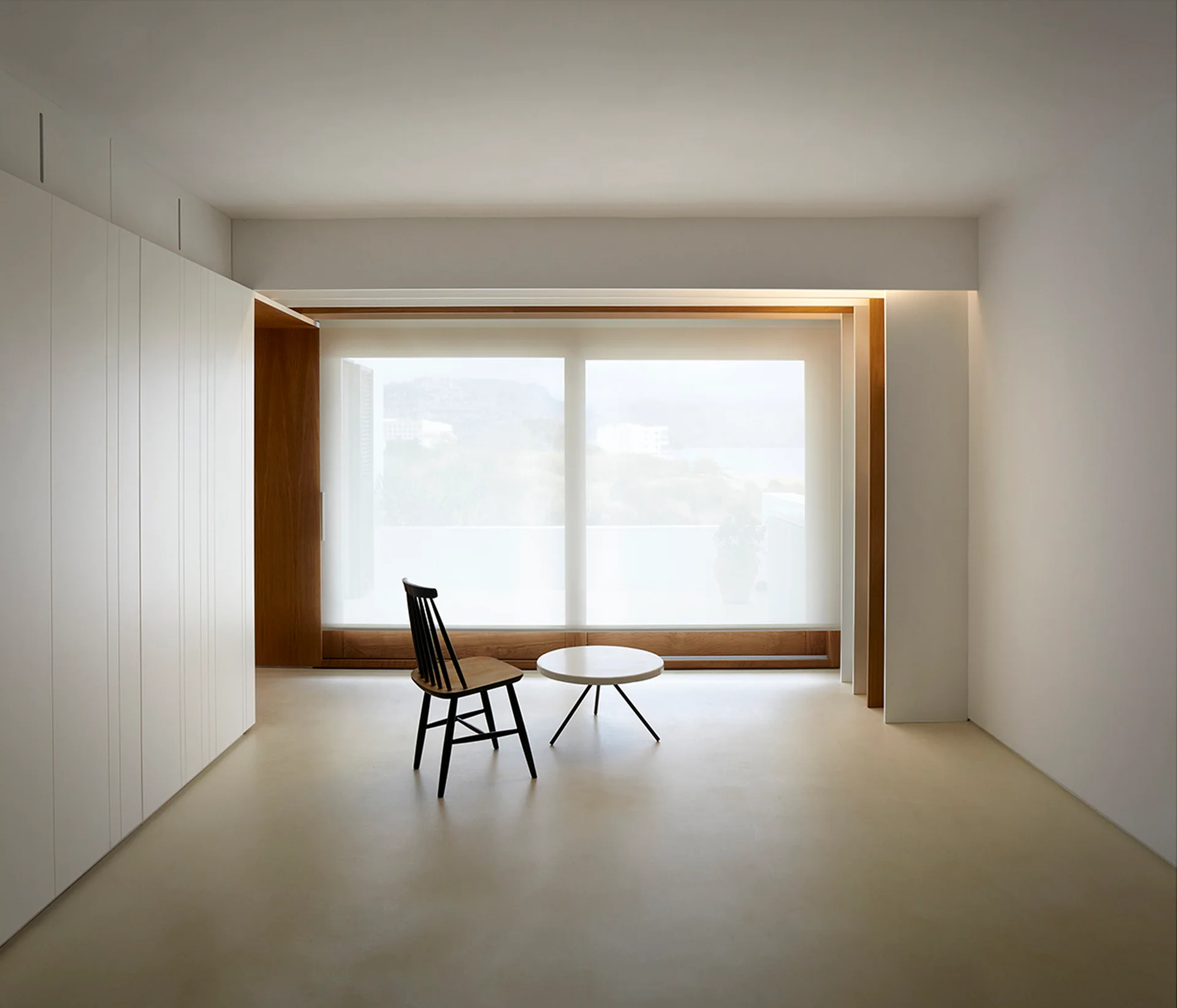 A minimalist white room features a black spindle chair, a small round table, floor-to-ceiling built-in cabinets, and a large window with a sheer shade.