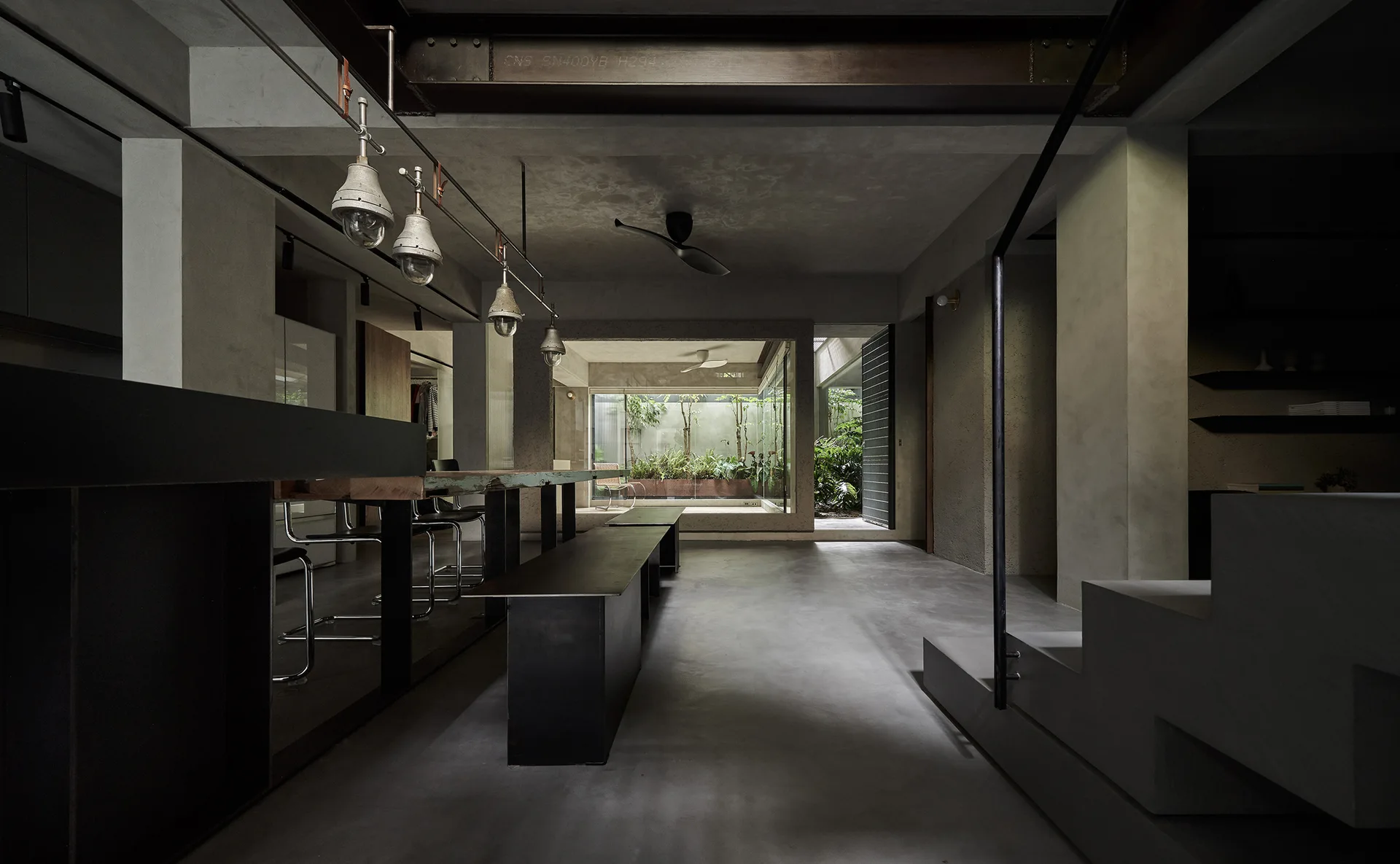 A minimalist gray concrete dining room featuring a long table, industrial pendant lights, and a view into a lush green courtyard.
