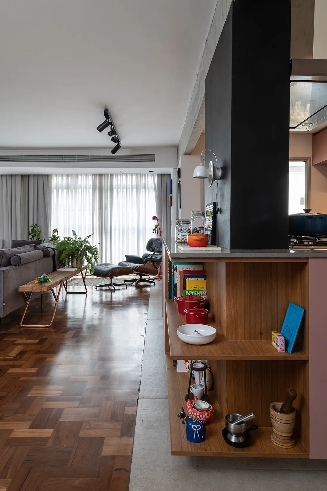 An open-plan interior showing wooden kitchen shelving on a black pillar beside a living area with a sofa, lounge chair, and herringbone parquet floor.