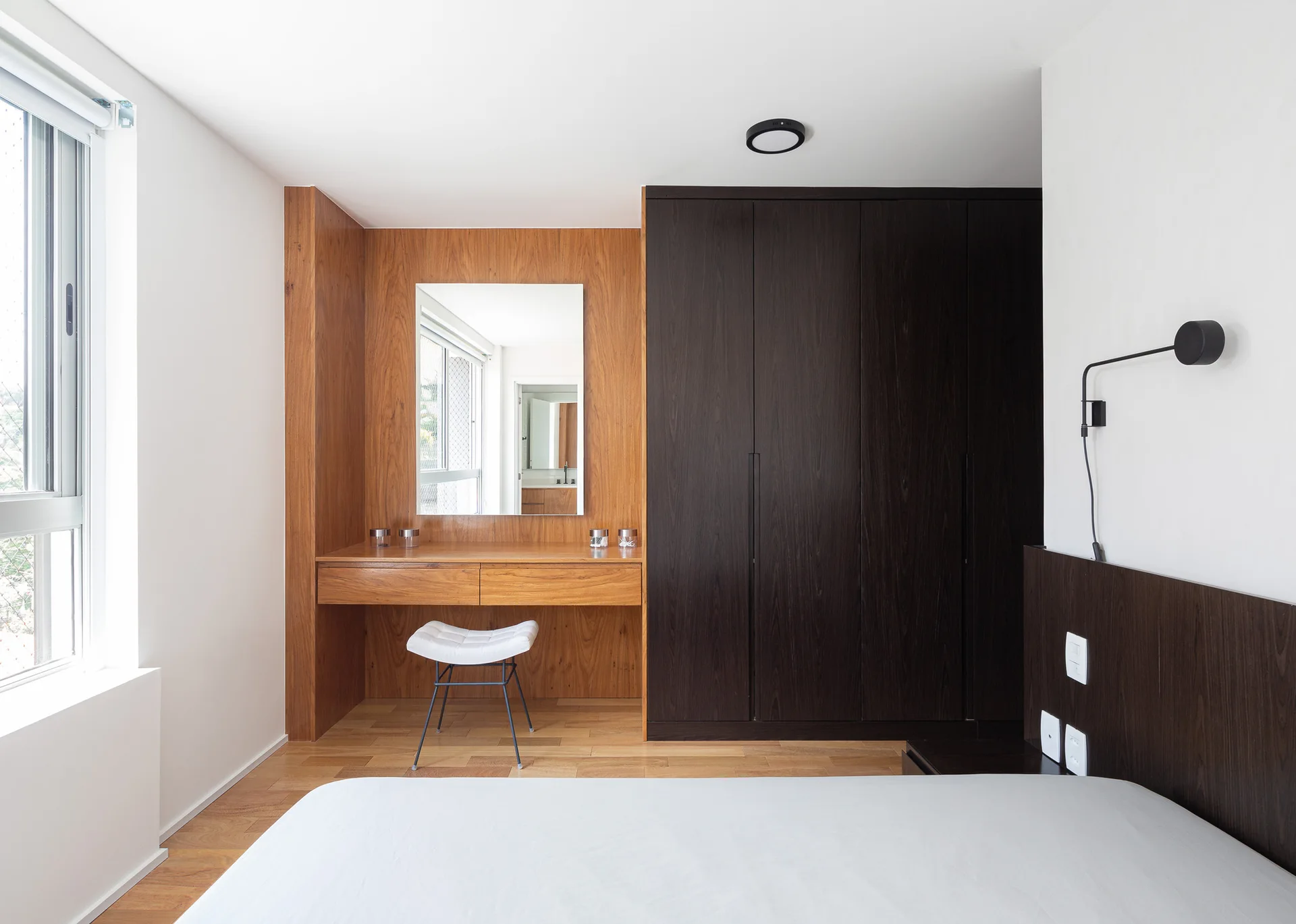 A modern bedroom featuring a built-in light wood vanity next to a tall dark wood wardrobe, with a white bed in the foreground.