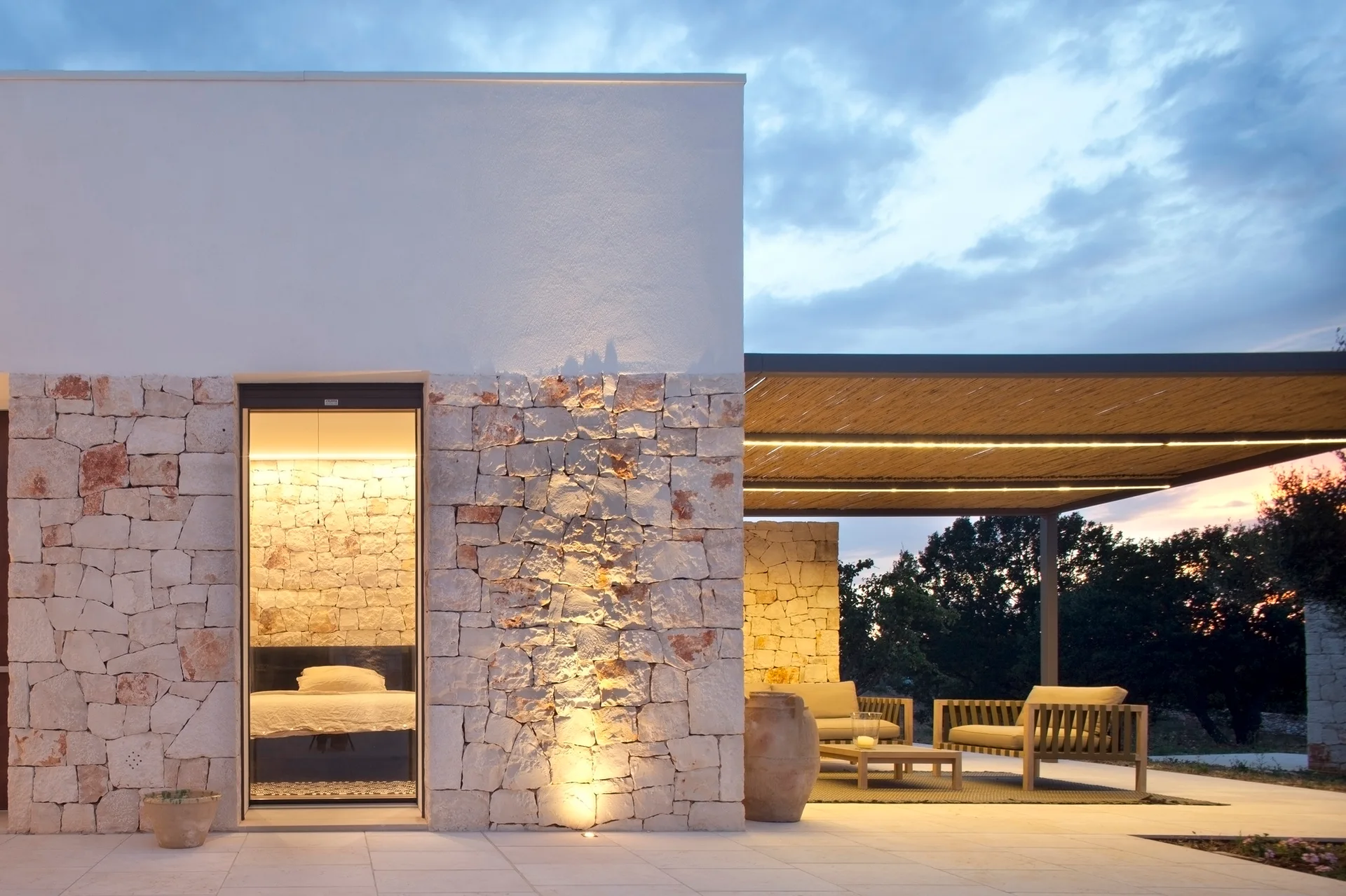 A modern outdoor patio at dusk features a slatted wood pergola over wooden armchairs, a dry stack stone facade, and a tall glass door.