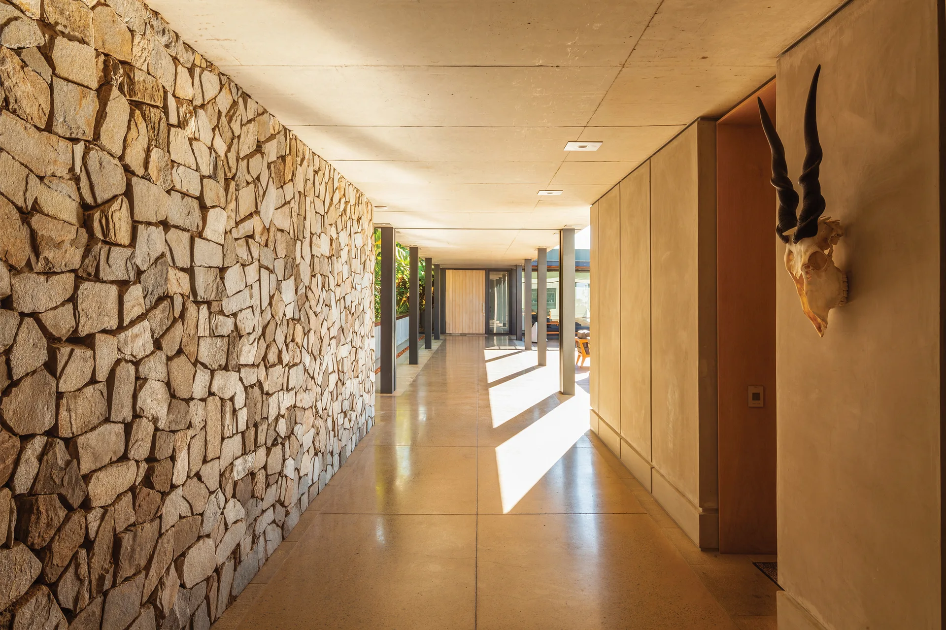 A long modern hallway featuring a rustic stone masonry wall, smooth concrete ceiling, polished floors, and a horned skull hanging on a side wall.