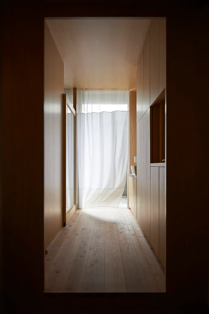 A narrow hallway with light wood paneling on the walls and ceiling, a plank floor, built-in storage, and a sheer white curtain at the end.