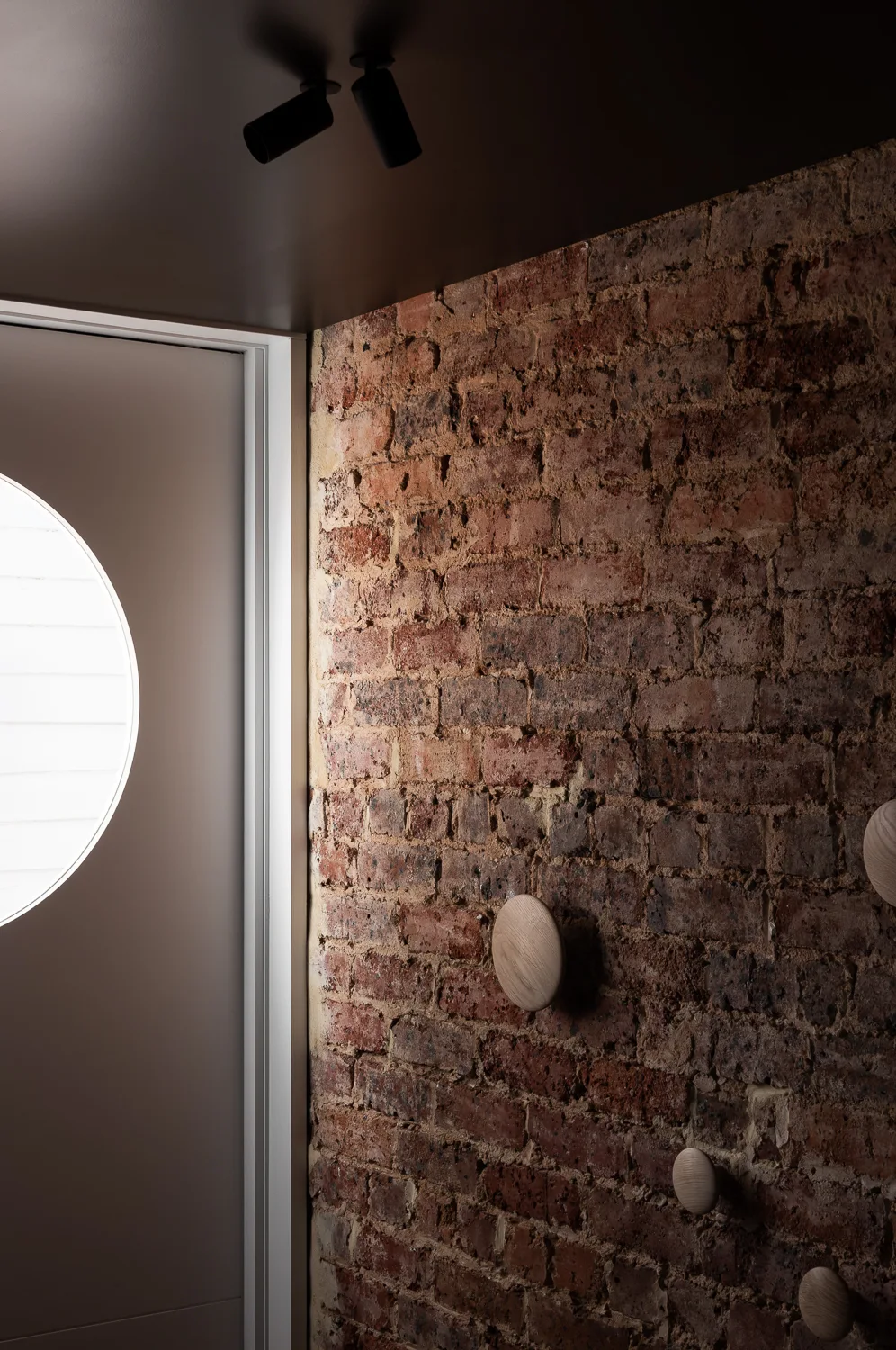An entryway features an exposed red brick wall with round wooden hooks next to a white wall with a circular window.