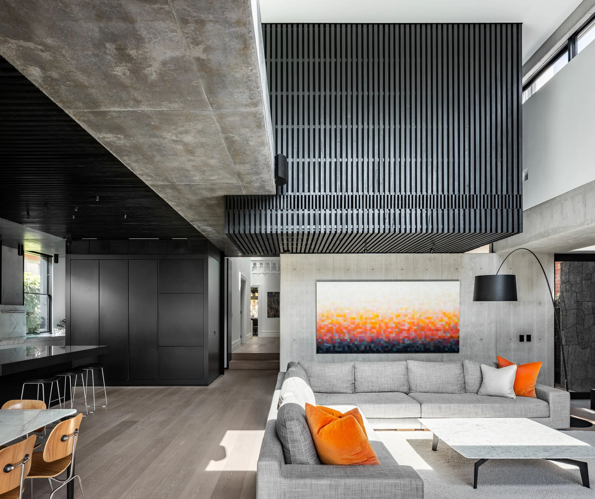 A modern living room features a grey sectional sofa with orange pillows under a black slatted feature wall and an exposed raw concrete beam.