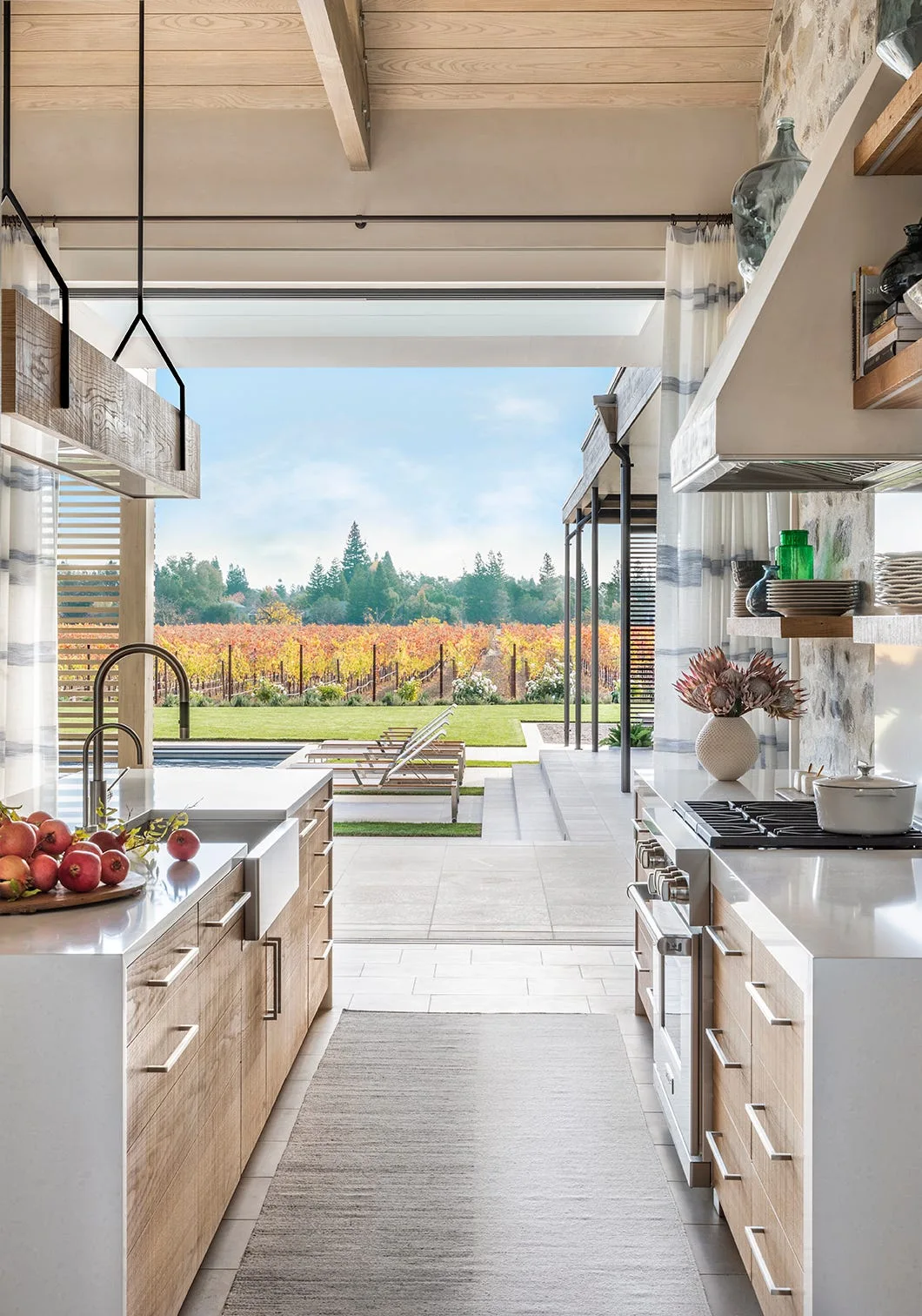 A modern farmhouse kitchen featuring light wood cabinets, white countertops, and a stone wall, looking out to an outdoor pool and vineyard.