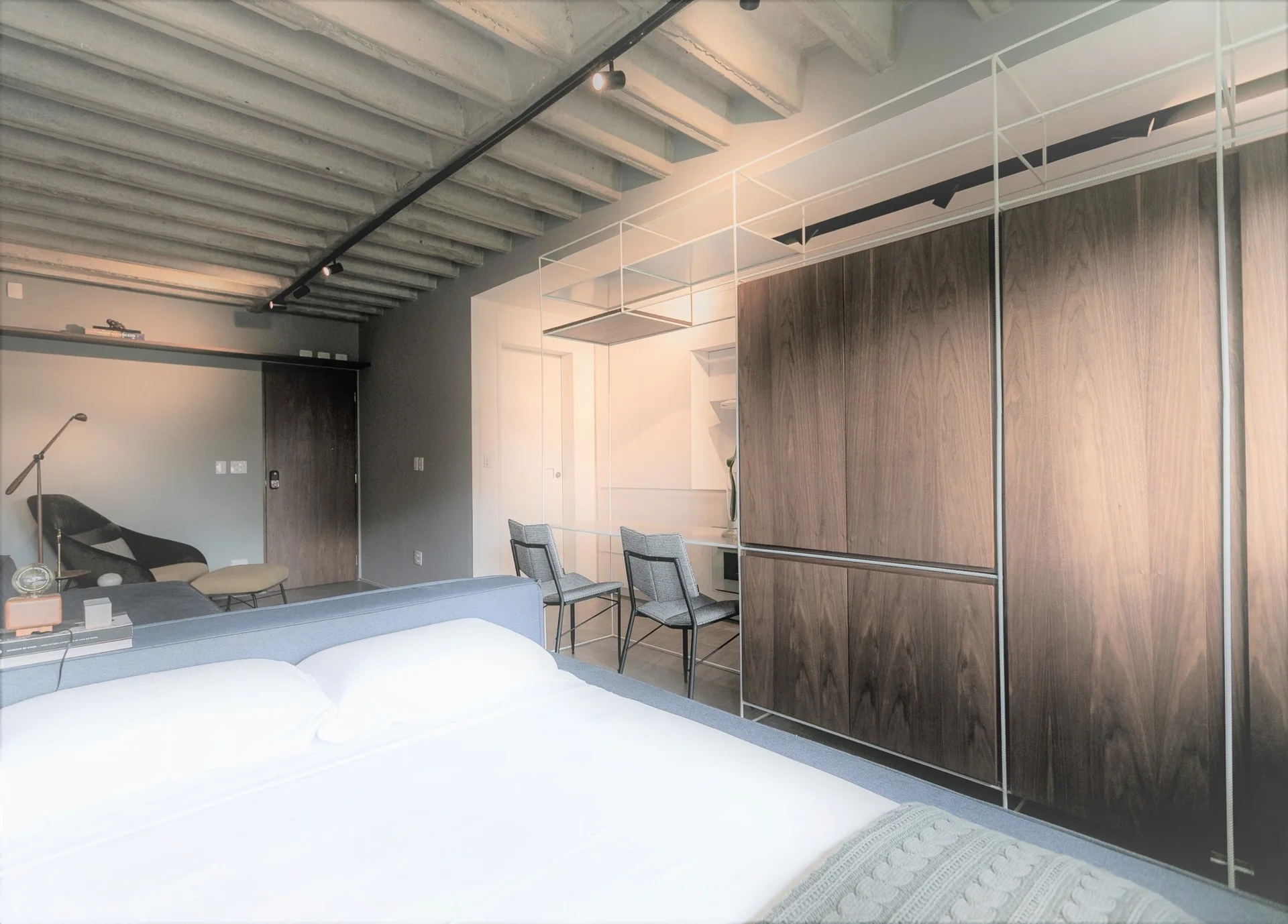 A modern bedroom with an exposed concrete joist ceiling, a blue platform bed, and built-in dark wood cabinetry containing an integrated desk.