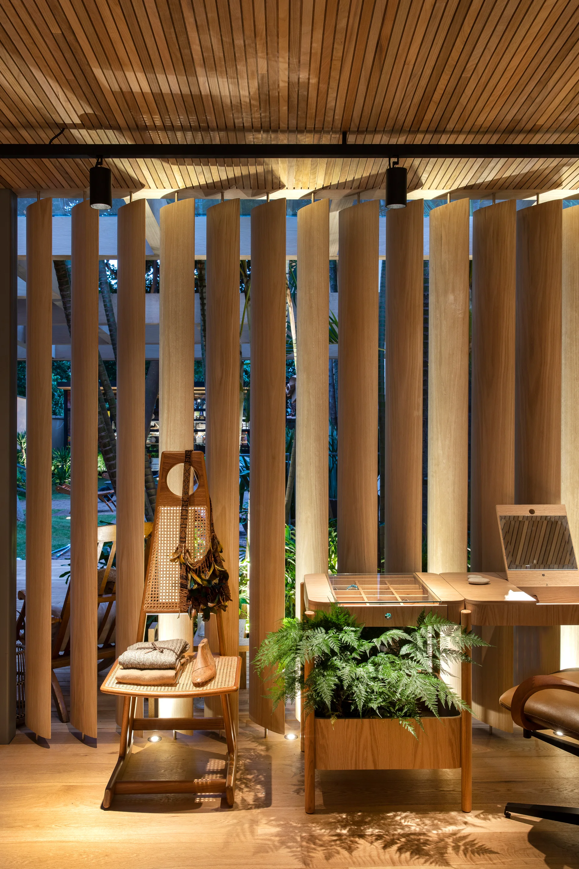 A vertical wood slat room divider stands behind a wooden vanity desk with an integrated fern planter and a cane-webbed accent chair.