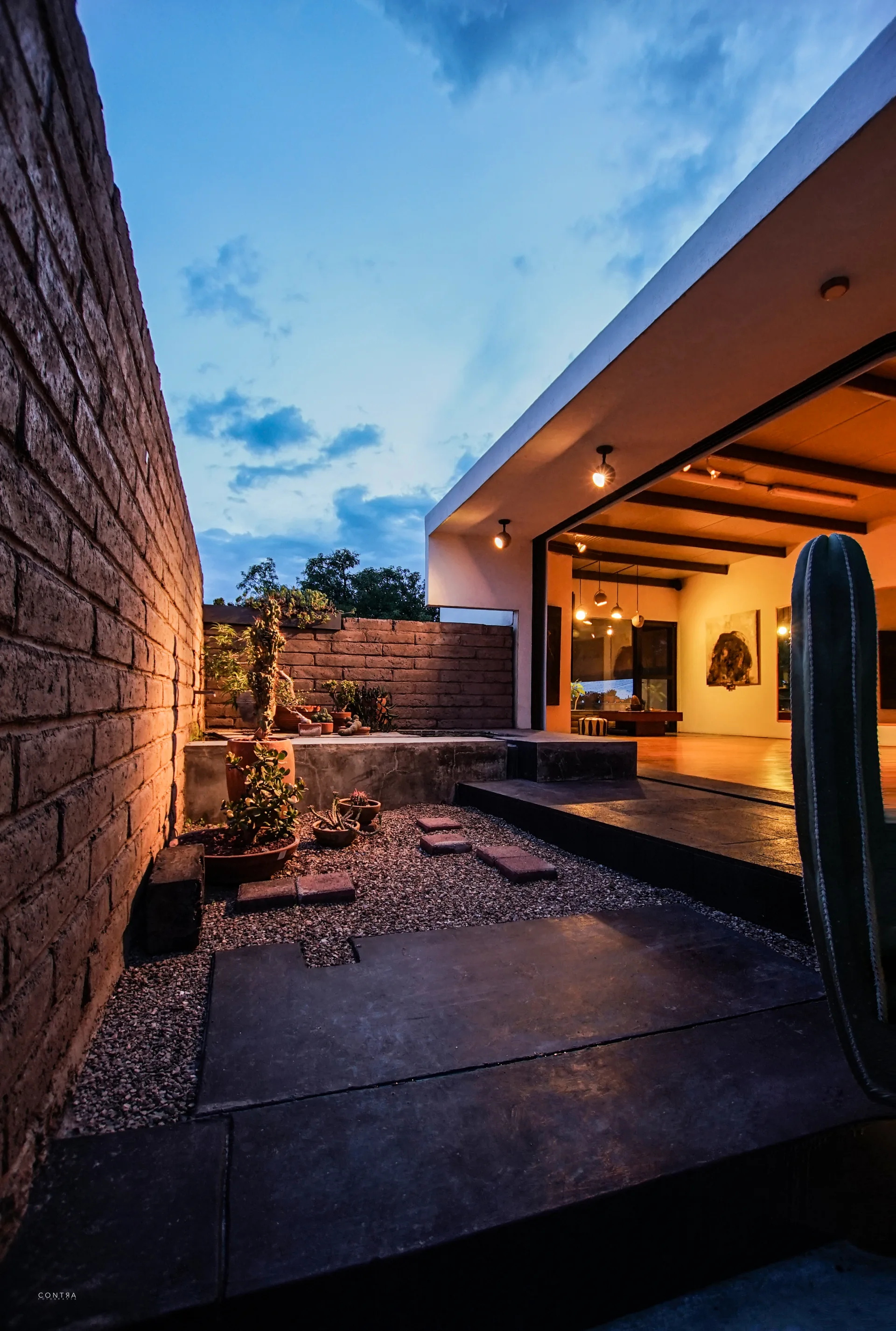 A modern courtyard with a textured brick wall, gravel path, and potted plants opens to a warm-lit interior room at dusk.