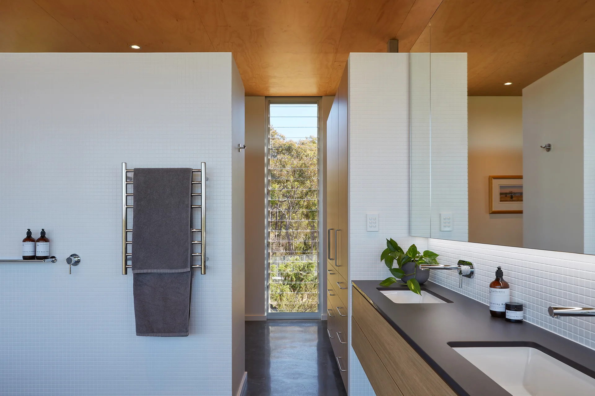A modern bathroom featuring a wood-front double vanity, white square mosaic tiled walls, a warm plywood ceiling, and a tall narrow louvred window.