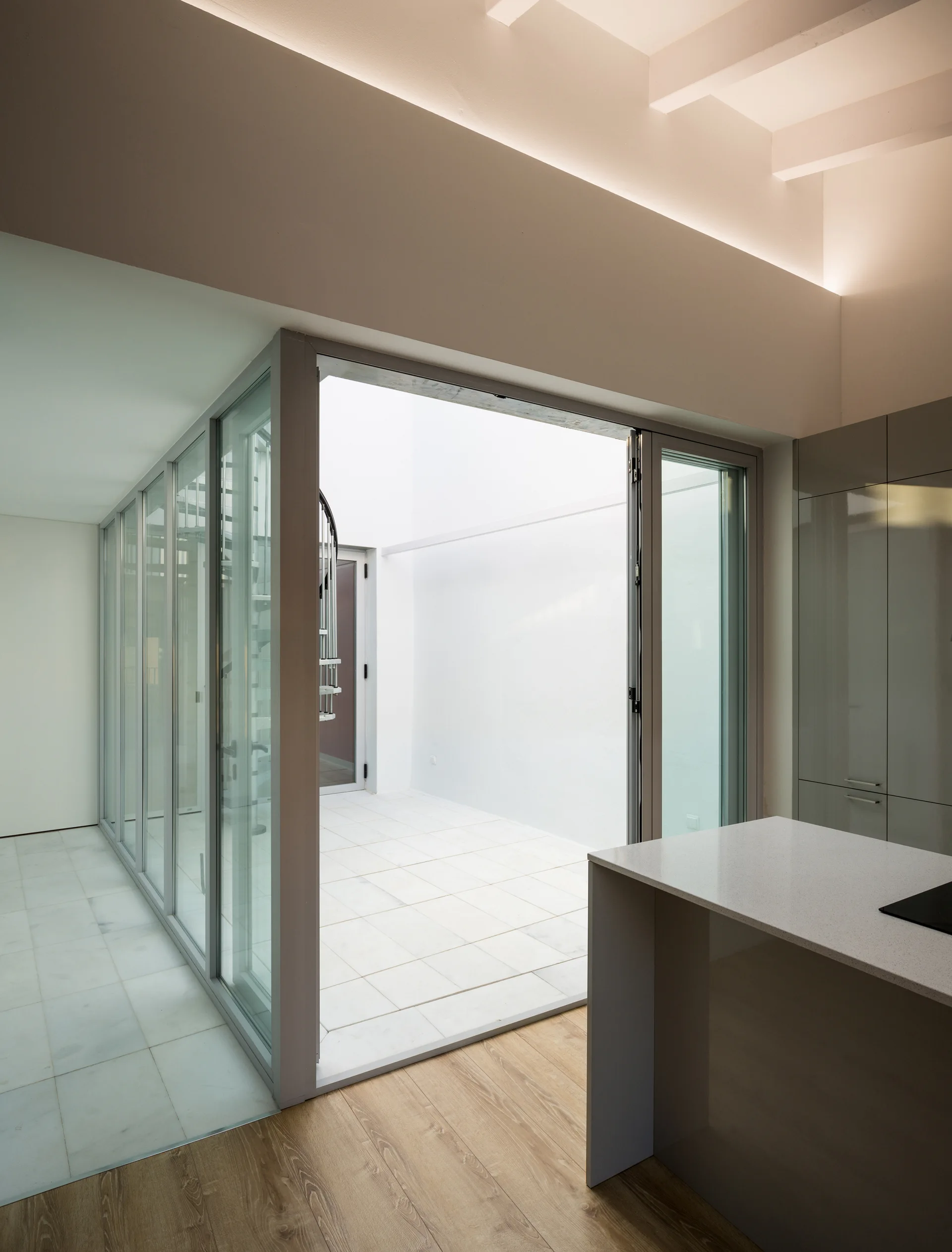 A minimalist kitchen with grey cabinetry opens to a glass-walled inner courtyard containing a white spiral staircase.