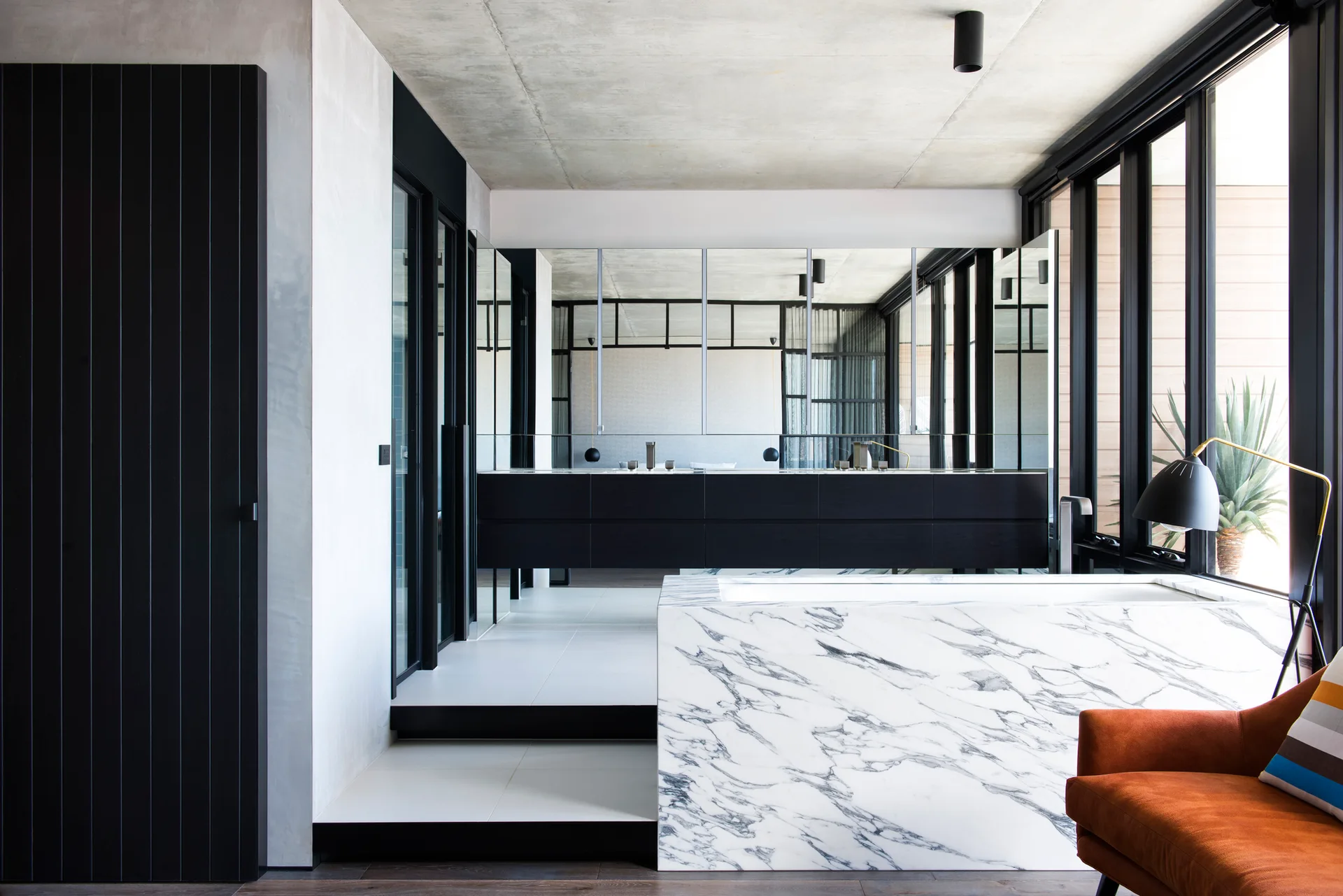 A modern bathroom features a rectangular marble-clad bathtub, a floating black vanity, a large wall mirror, and an exposed concrete ceiling.