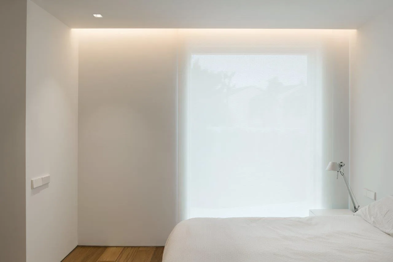 A minimalist white bedroom with wood flooring, a simple bed with a metal bedside lamp, and warm cove lighting along the ceiling edge.