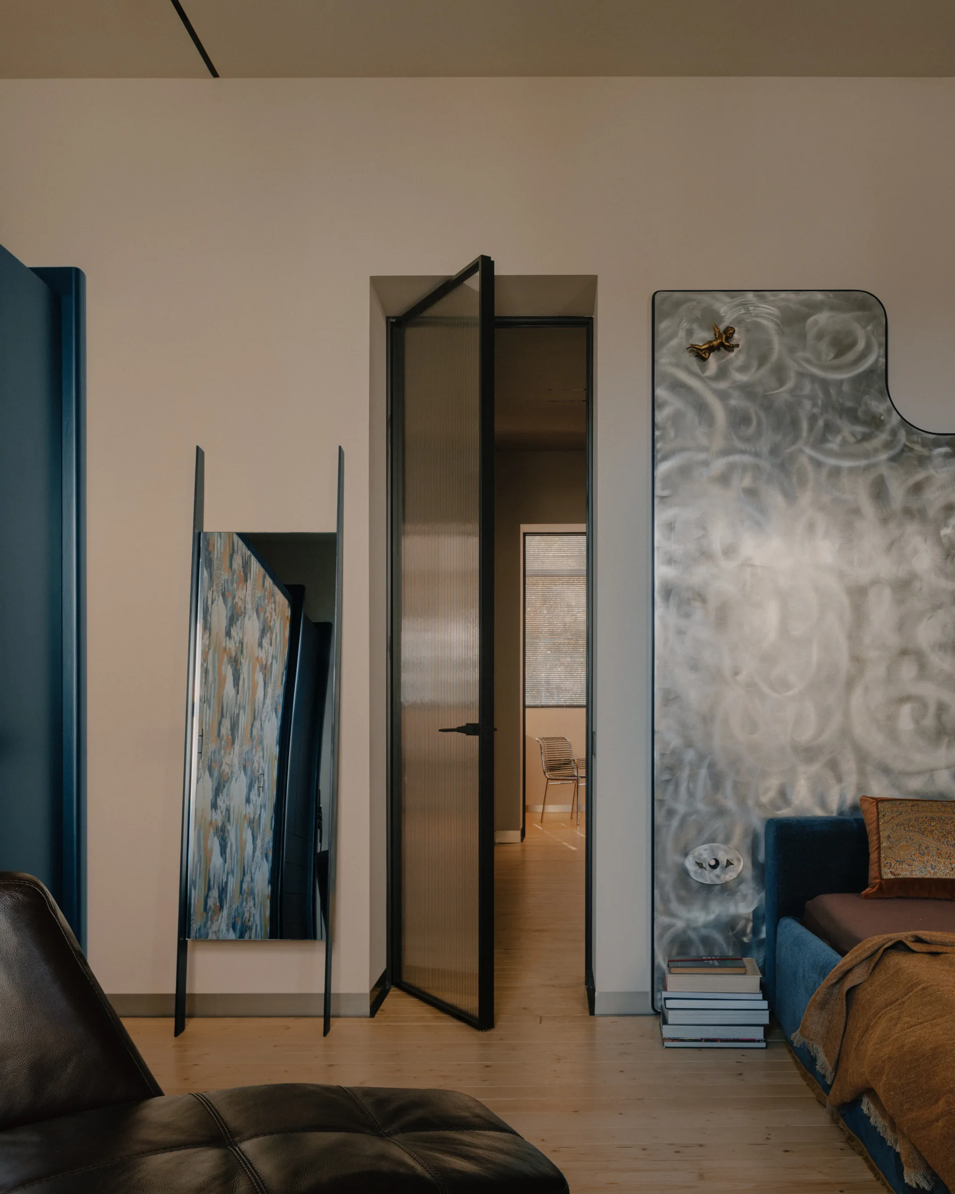 A modern bedroom features a black-framed fluted glass door, a blue bed, and a swirling textured silver metal panel on the wall.
