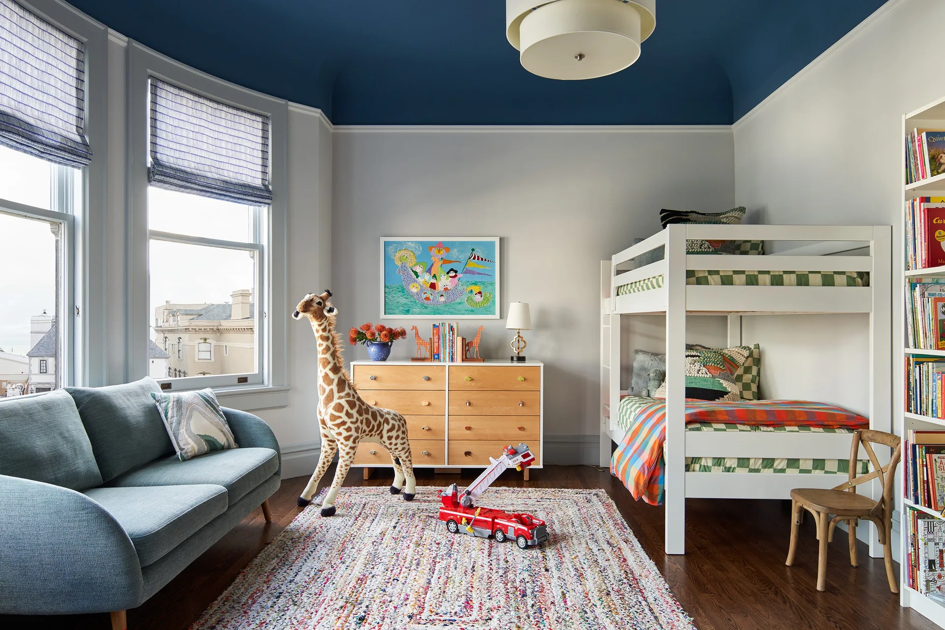 A kids' bedroom with a white bunk bed, a light blue sofa near a bay window, a light wood dresser, and a painted deep blue ceiling.