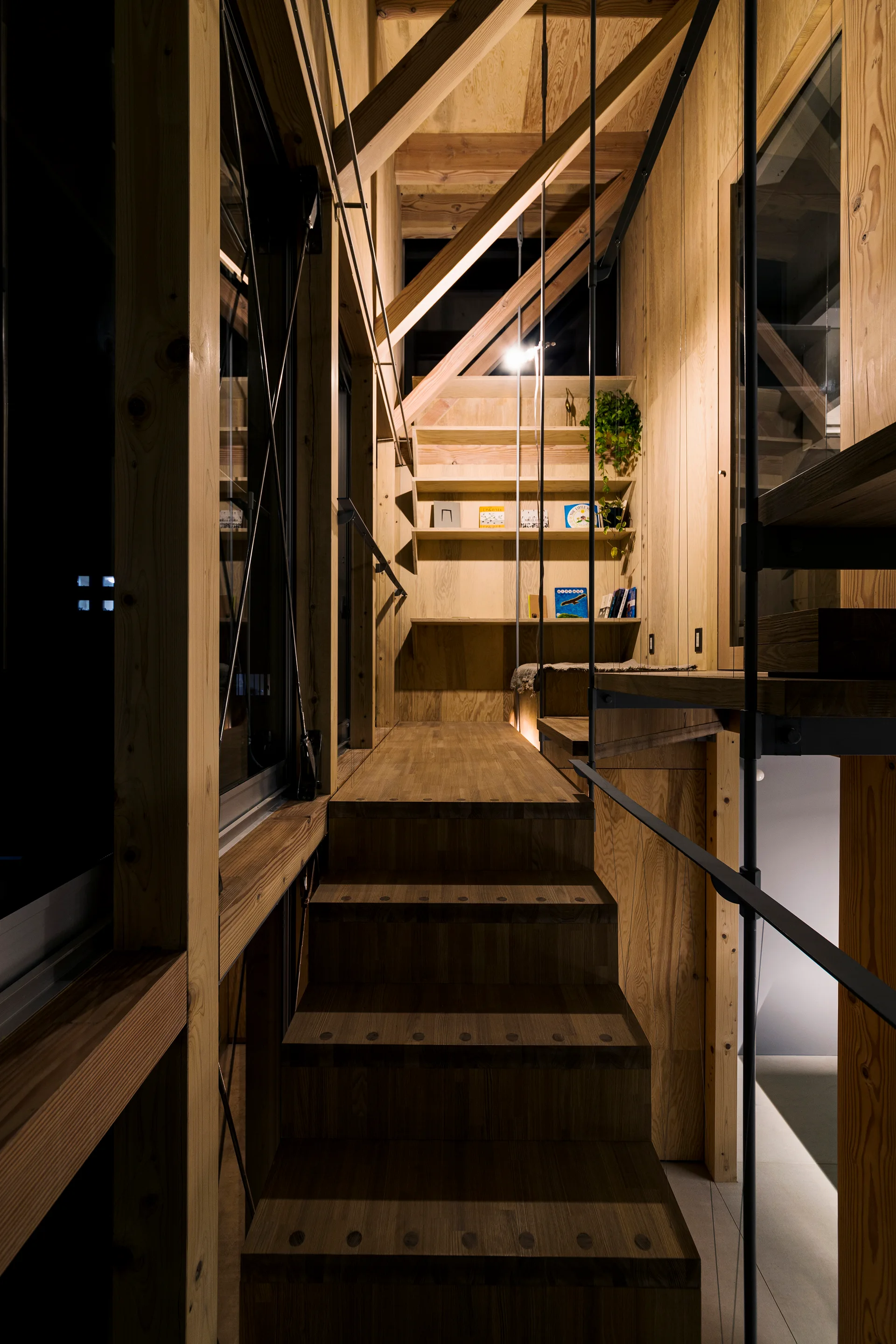 Wooden stairs ascend through a structure of exposed timber beams next to a built-in plywood bookshelf illuminated by warm spotlighting.