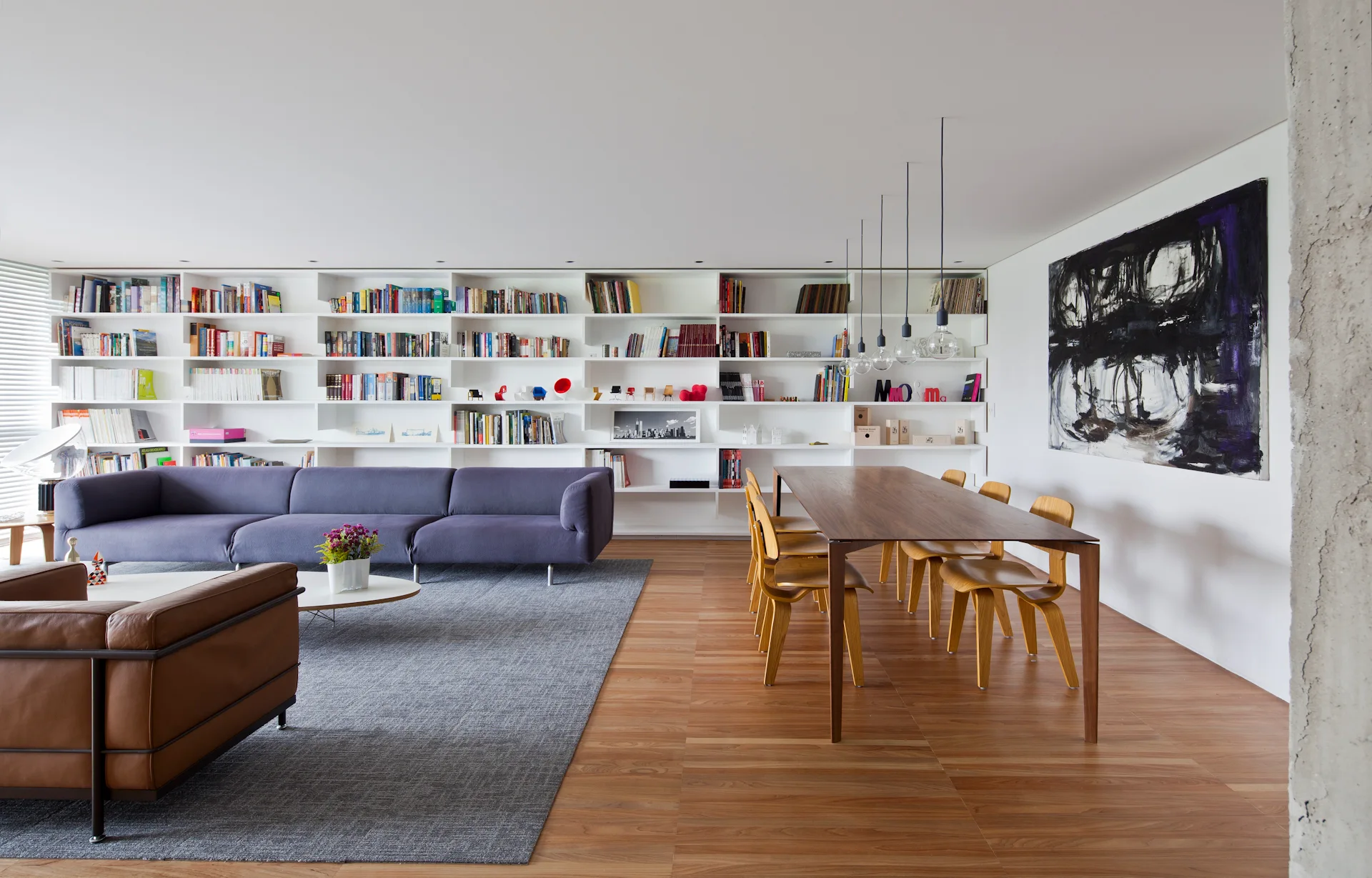 An open-plan living and dining area featuring a massive white built-in bookshelf, a purple sectional sofa, a wooden dining table, and hardwood floors.