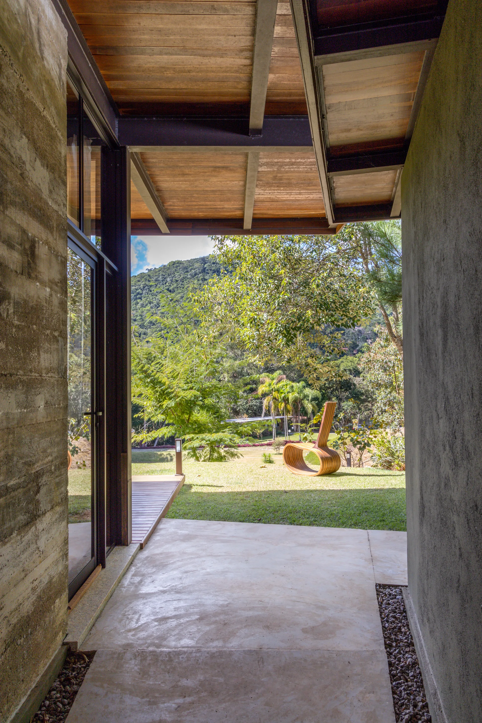 A concrete outdoor transition space with a wood ceiling leads to a grassy lawn featuring a sculptural wooden lounge chair against a forest background.