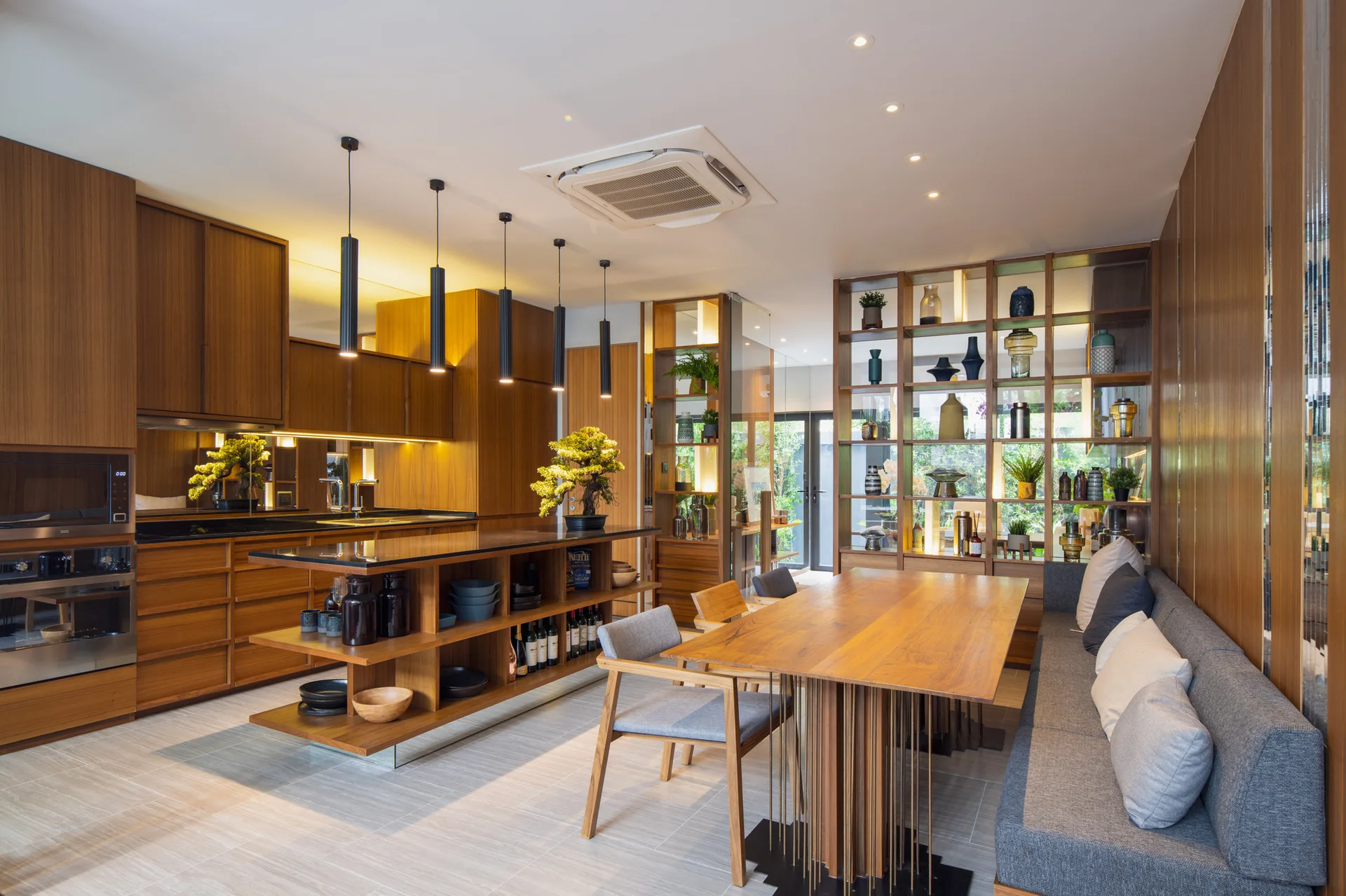 A modern wood kitchen and dining space with a central island, a wooden table with a gray upholstered banquette bench, and an open display shelving unit.