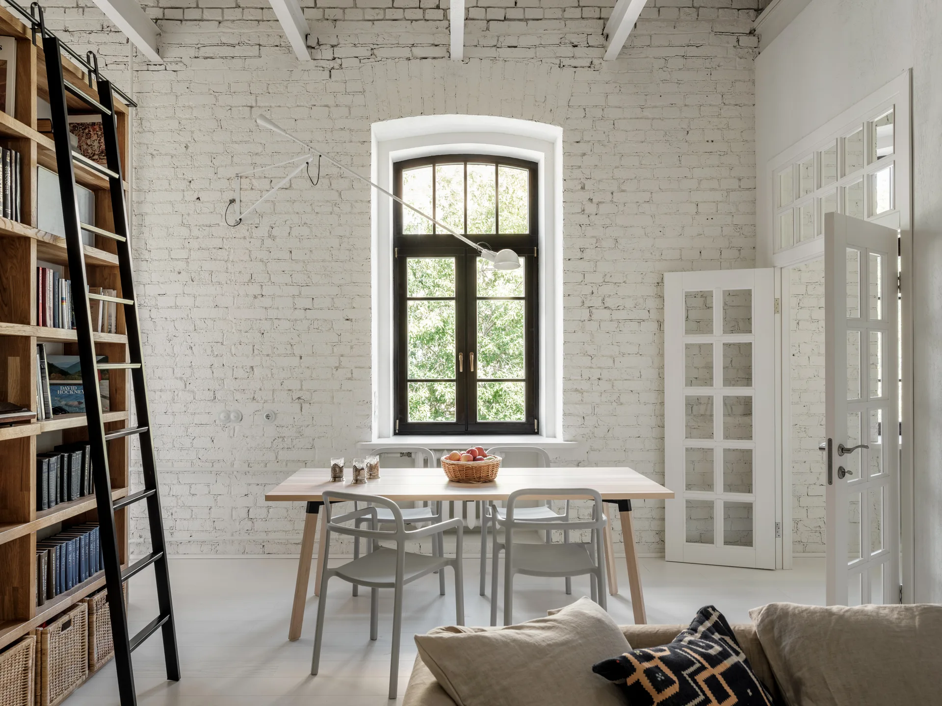 A light wood dining table with grey chairs sits in front of a tall arched window, set against a white painted brick wall with a wooden bookshelf.