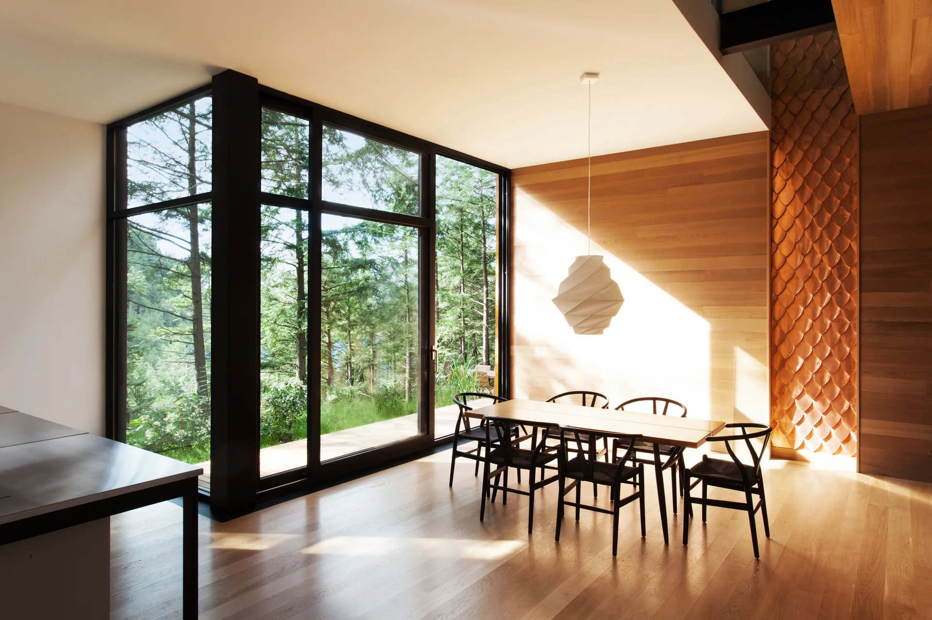 A modern dining room features a wooden table and black chairs set against floor-to-ceiling windows looking out onto a dense forest.