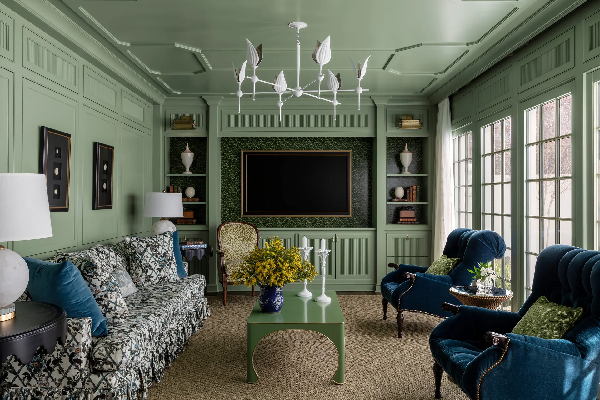 A sage green paneled living room features a matching coffered ceiling, a floral print sofa, and two deep blue velvet armchairs on a woven rug.