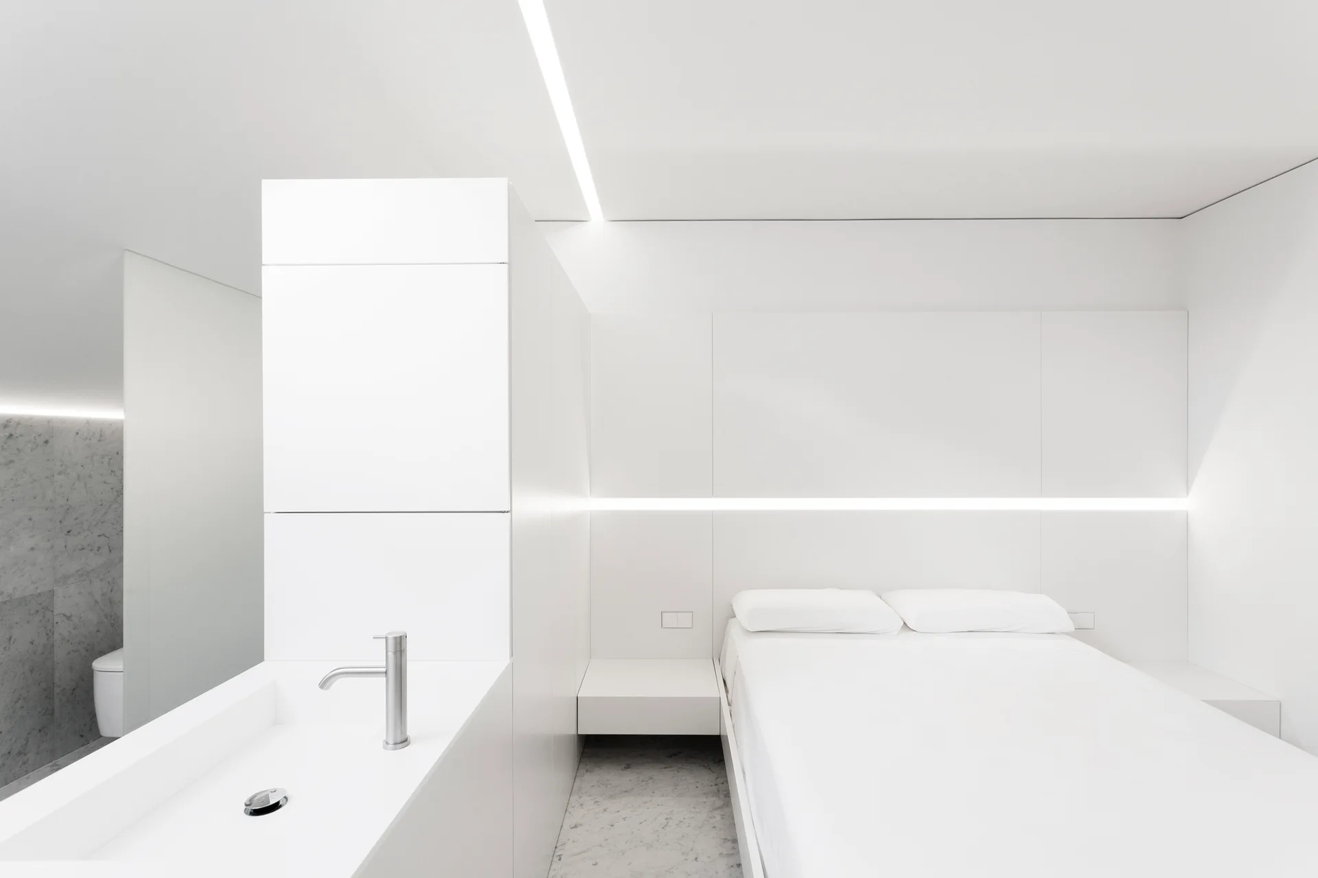 A minimalist white bedroom features a platform bed, integrated linear wall lighting, and a white partition with an integrated sink and chrome faucet.