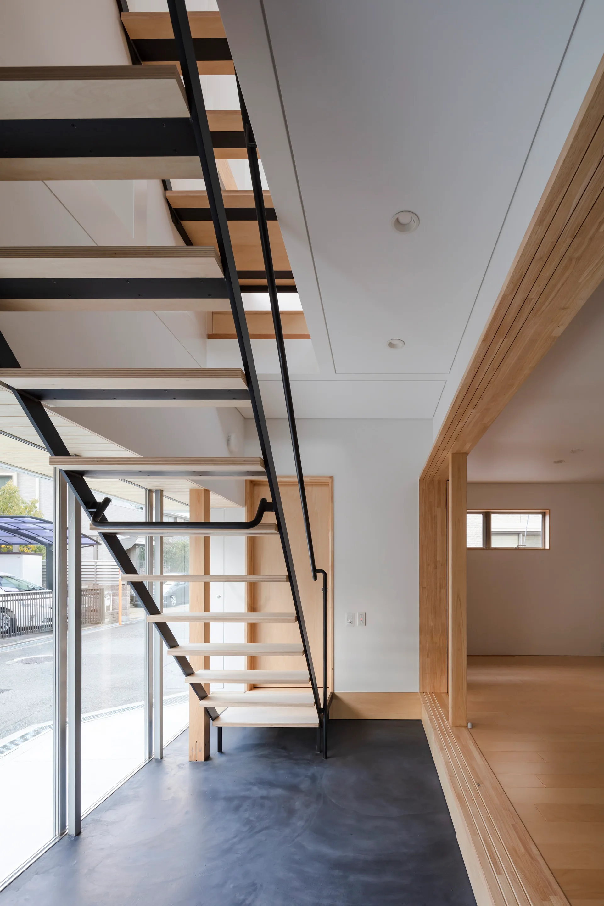 An open-riser wooden staircase with black metal framing stands on a dark concrete floor next to a tall window and a light wood partition.
