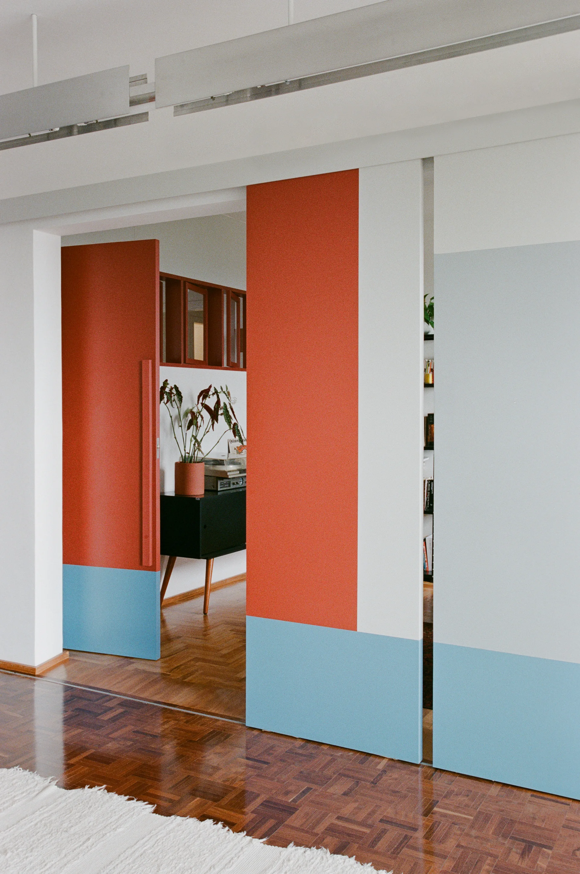 Color-blocked sliding doors in terracotta, white, and light blue open to reveal a living room with herringbone parquet floors and a black cabinet.